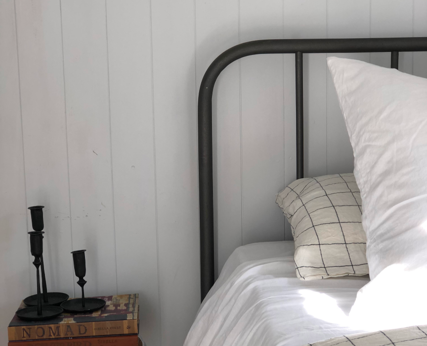 A minimalistic bedroom scene featuring a metal bed frame, white sheets with a grid-patterned pillow, and a bedside stack of books. Beside the stack are three black candle holders against a light-colored wooden wall background.