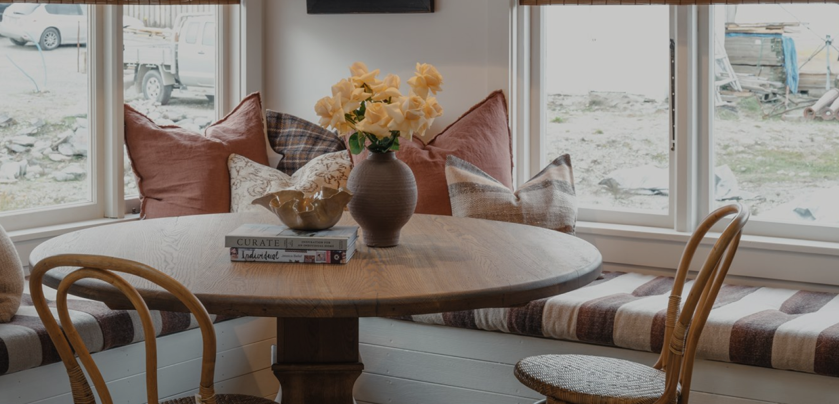 Cozy dining area with a round wooden table, surrounded by wicker chairs and a cushioned bench. A vase with yellow flowers and a stack of books are on the table. Large windows provide natural light and an outdoor view.