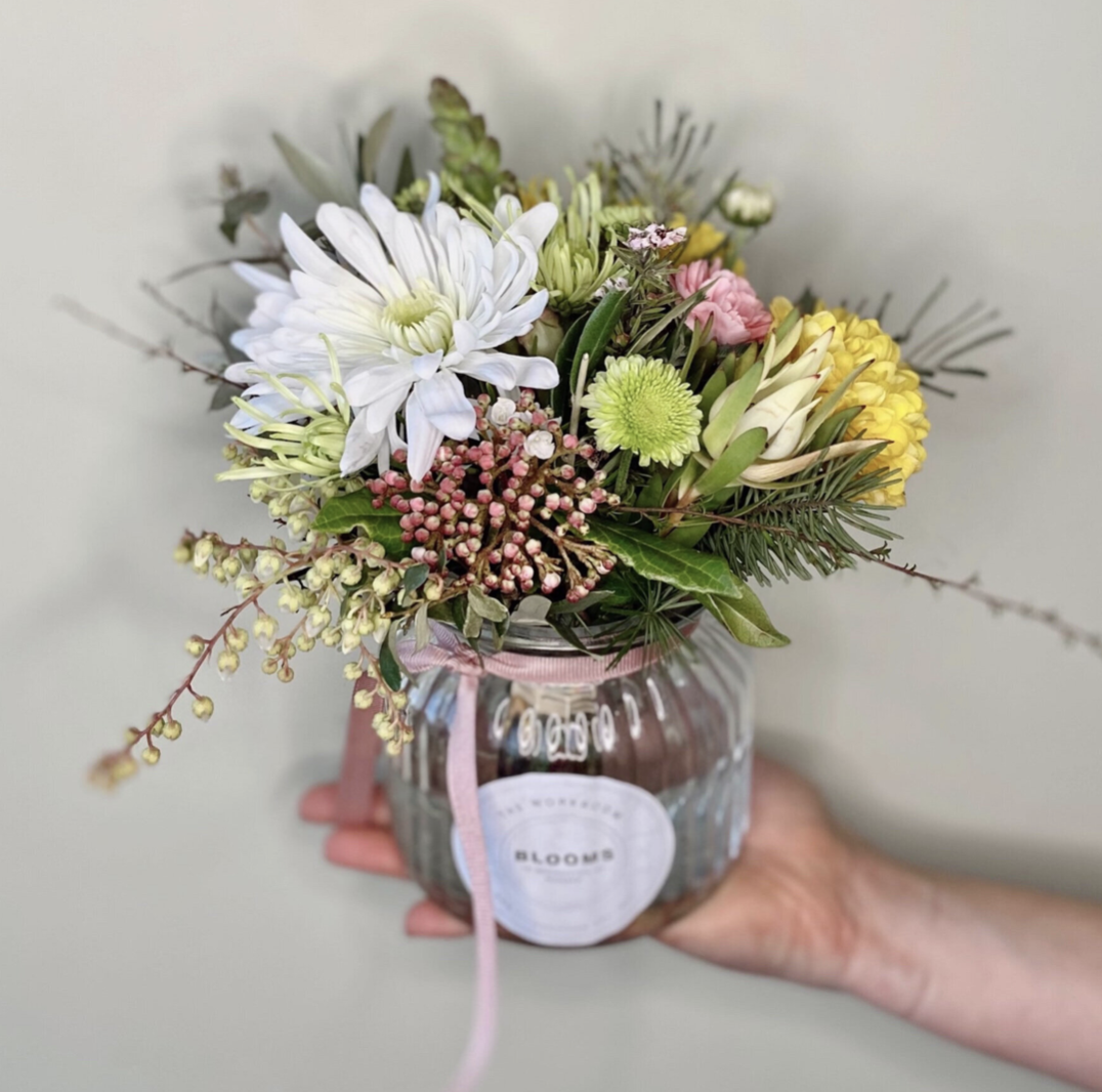 A person holds a POSY JAR by The Workroom, featuring a small bouquet of assorted flowers, including white daisies and pink roses, elegantly arranged in a clear jar with a pink ribbon. The background is plain and light-colored.