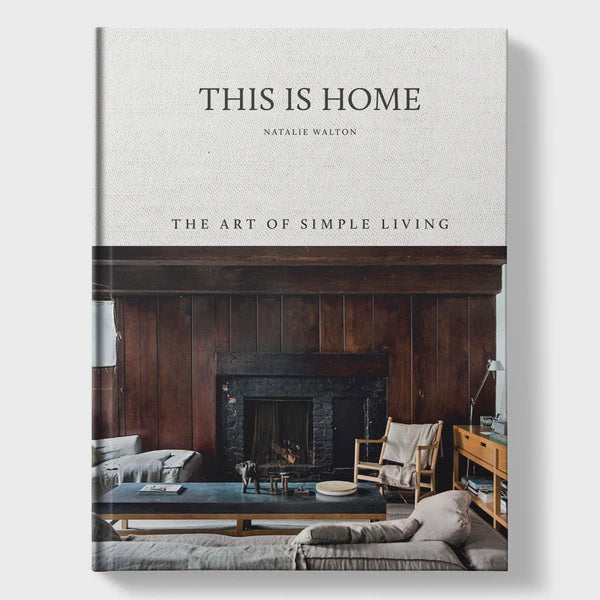 Book cover titled THIS IS HOME - NATALIE WALTON by Nation Wide Books. It showcases a cozy living room featuring a wood-paneled fireplace, sofas, an armchair, and wooden furniture, all beautifully captured to evoke comfort and minimalism in authentic homes.