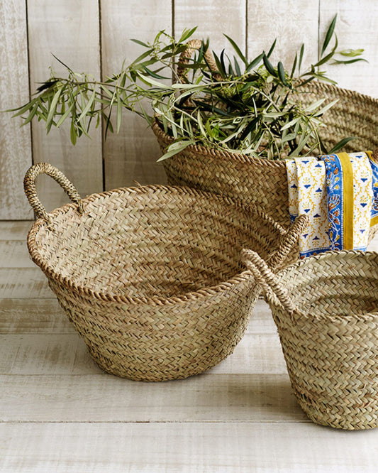 Three SOUK MARKET BASKETS by Epicure Trader in varying sizes rest on a wooden floor. The largest basket, expertly crafted as a multipurpose basket, contains green branches. A cloth with a blue and yellow pattern drapes over its edge. In the background, a wooden wall completes the rustic scene.