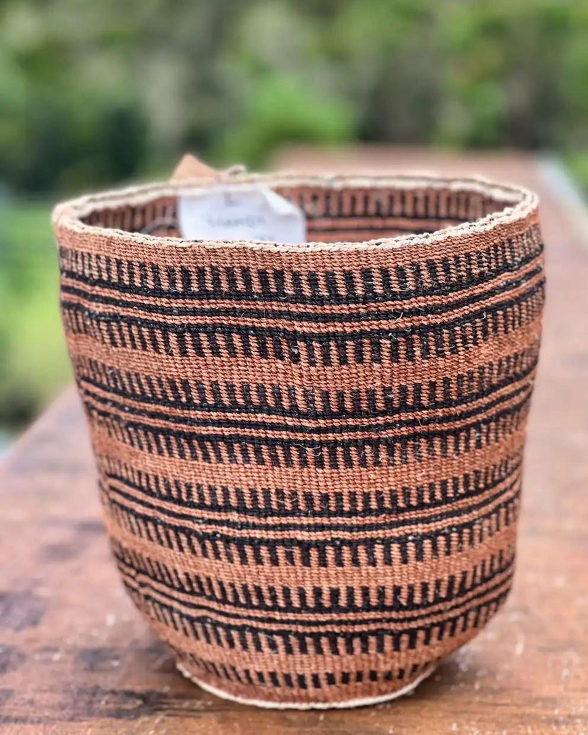 The SUBIRA BASKET by Manabaskets rests on a wooden surface, featuring its black and brown striped patterns that highlight the craftsmanship of women artisans in Kenya. The background is softly blurred with hints of greenery, creating a natural setting. A small label on the basket's rim indicates its fair trade status.