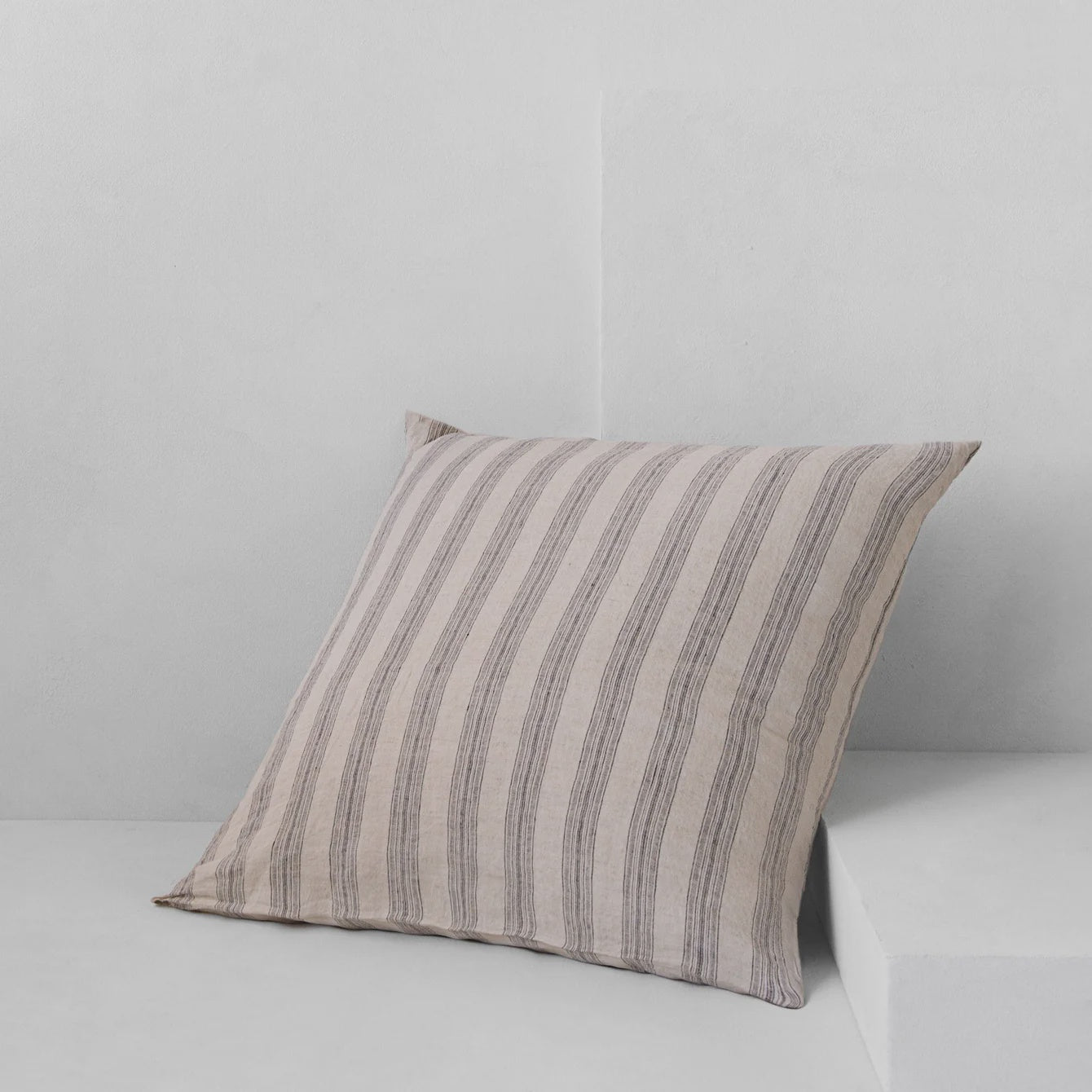 On a light gray surface, a HALE BASIX STRIPE EURO PILLOWCASE by Hale Mercantile Co, featuring vertical gray and beige stripes, is propped against a matching wall backdrop. The arrangement exudes a minimalistic and modern aesthetic reminiscent of sophisticated European pillowcase designs.