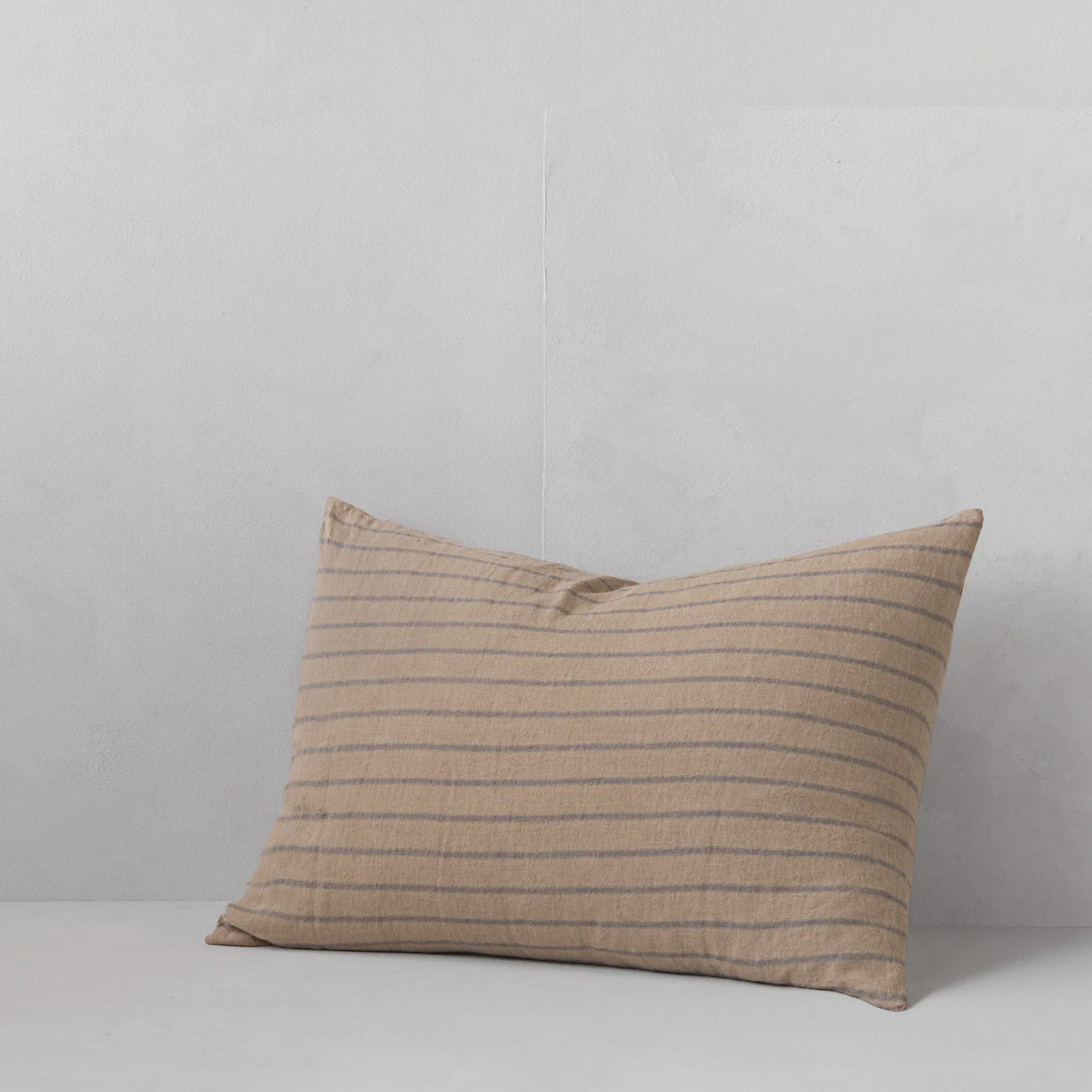 A HALE BASIX STRIPE PILLOWCASE by Hale Mercantile Co in beige featuring thin brown horizontal stripes sits on a light gray surface against a matching gray background. Made from pure linen, the pillowcase is slightly crumpled, offering a casual and relaxed appearance.
