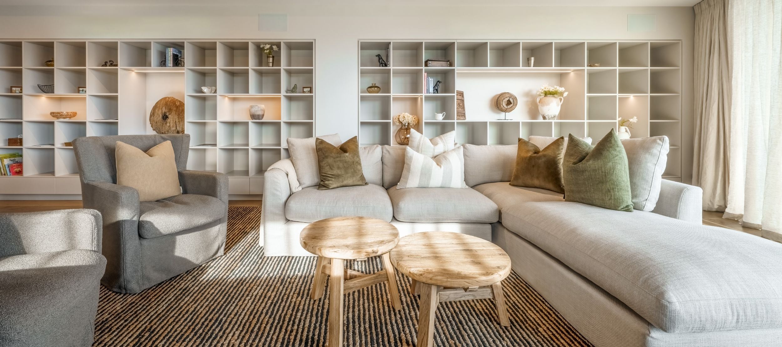A cozy living room featuring a light gray sectional sofa with green and beige cushions, a gray armchair, and two round wooden coffee tables. The background showcases a wall of bookshelves filled with decorative items, lit by natural sunlight.