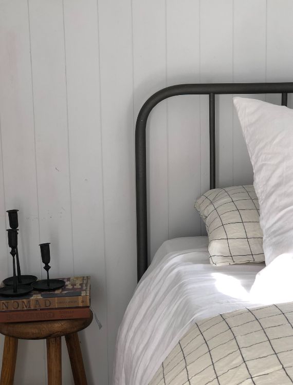 A cozy bedroom scene with a metal-framed bed featuring white and checkered bedding. To the left, a wooden side table holds two black candlesticks and a book. The background wall is painted white, creating a serene atmosphere.