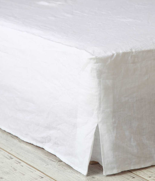 In a corner view, the MM Linen Valance from MM Linen elegantly drapes over a bed or table, extending to the floor. Made from pure linen, it features a minimalist design with a refined slit at the corner and textured appeal, perfectly complementing the light wood floor.