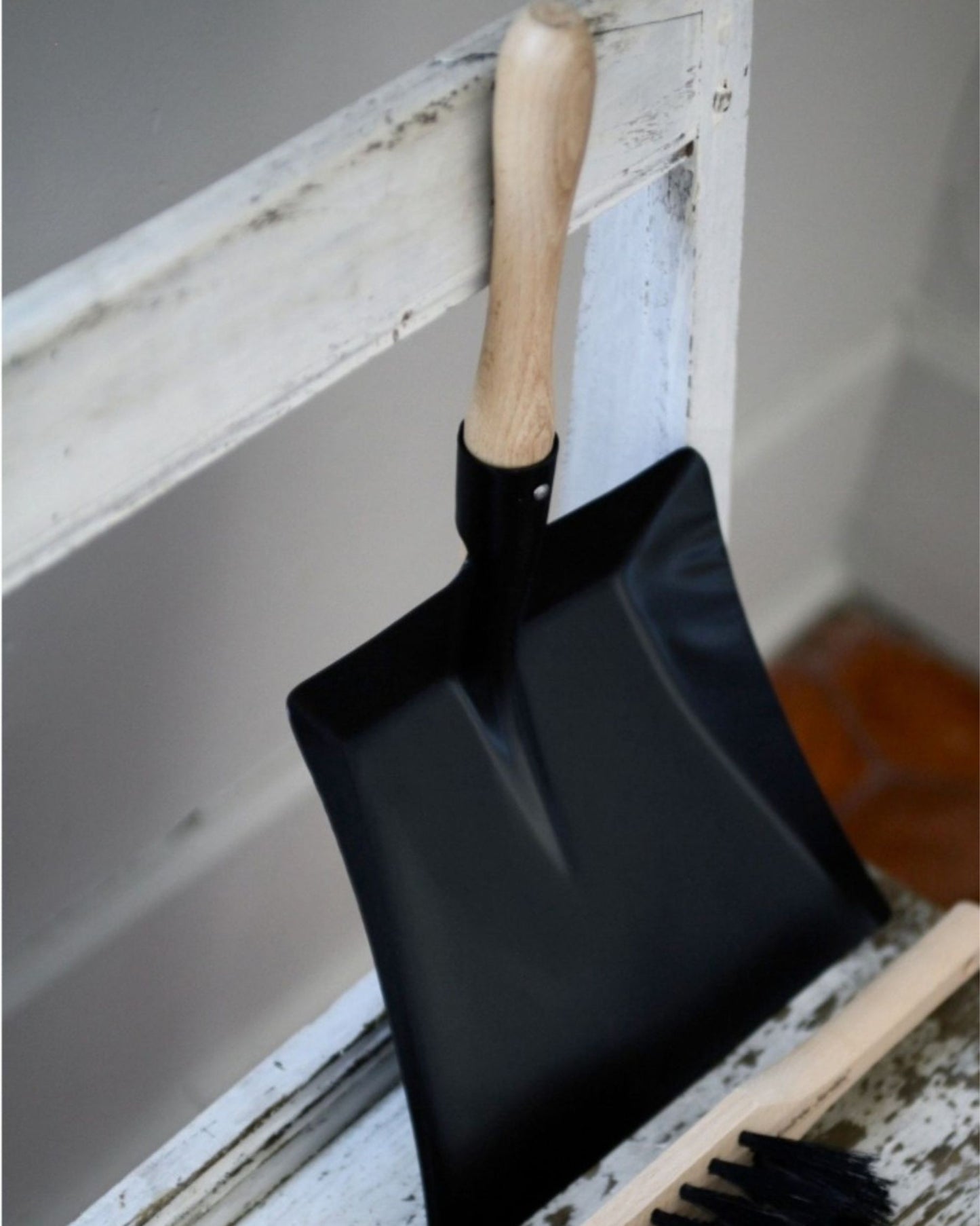 A black DUSTPAN with a French beechwood handle by Andree Jardin hangs gracefully on a white wooden frame. In the foreground, a petite brush lies on the wooden surface, highlighted by the warm tone of terracotta tiles in the background.