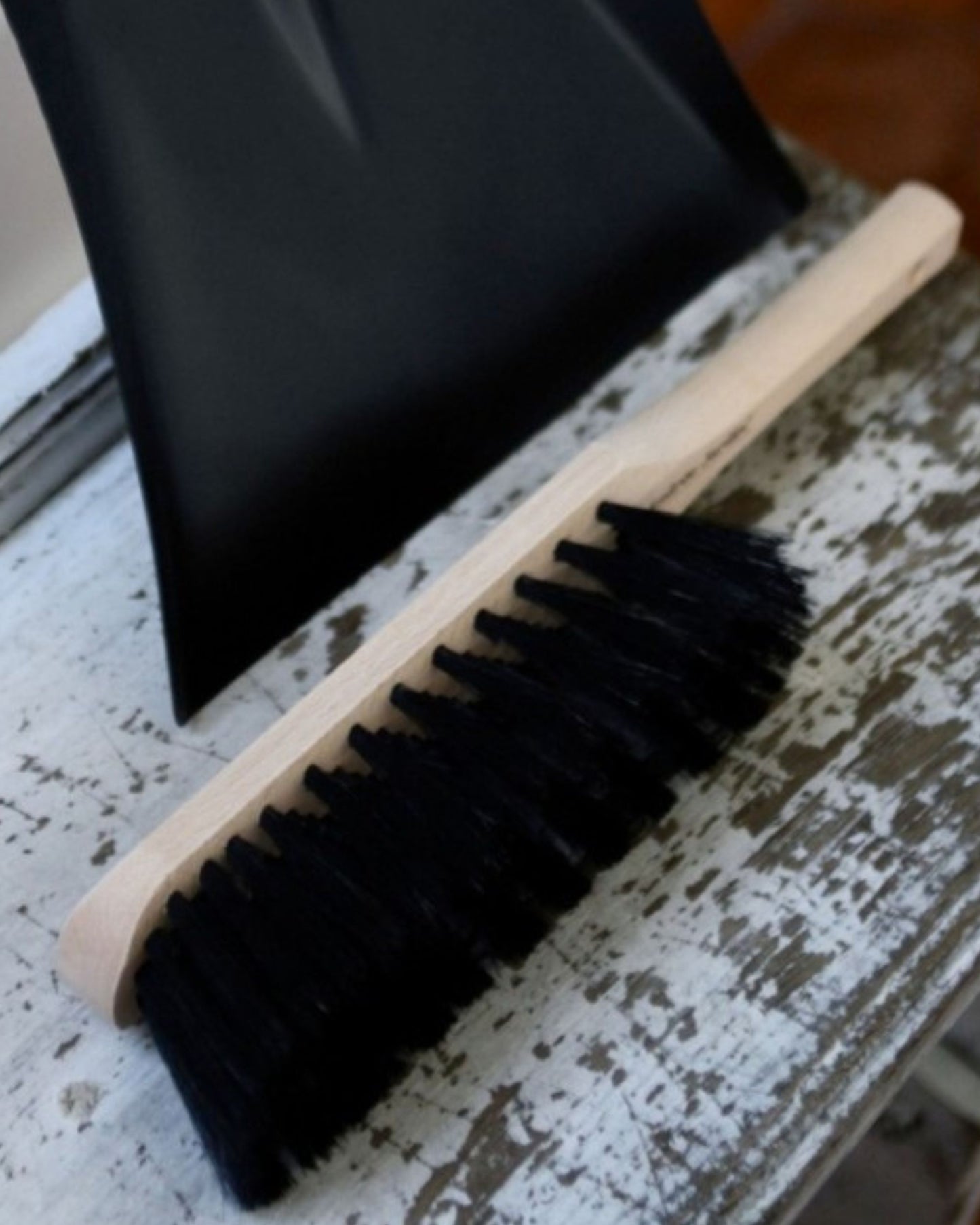 A handcrafted HAND BRUSH by Andree Jardin, featuring a wooden handle and natural bristles, rests on a weathered, white-painted wooden surface. Nearby, a black dustpan is partially visible in the background.