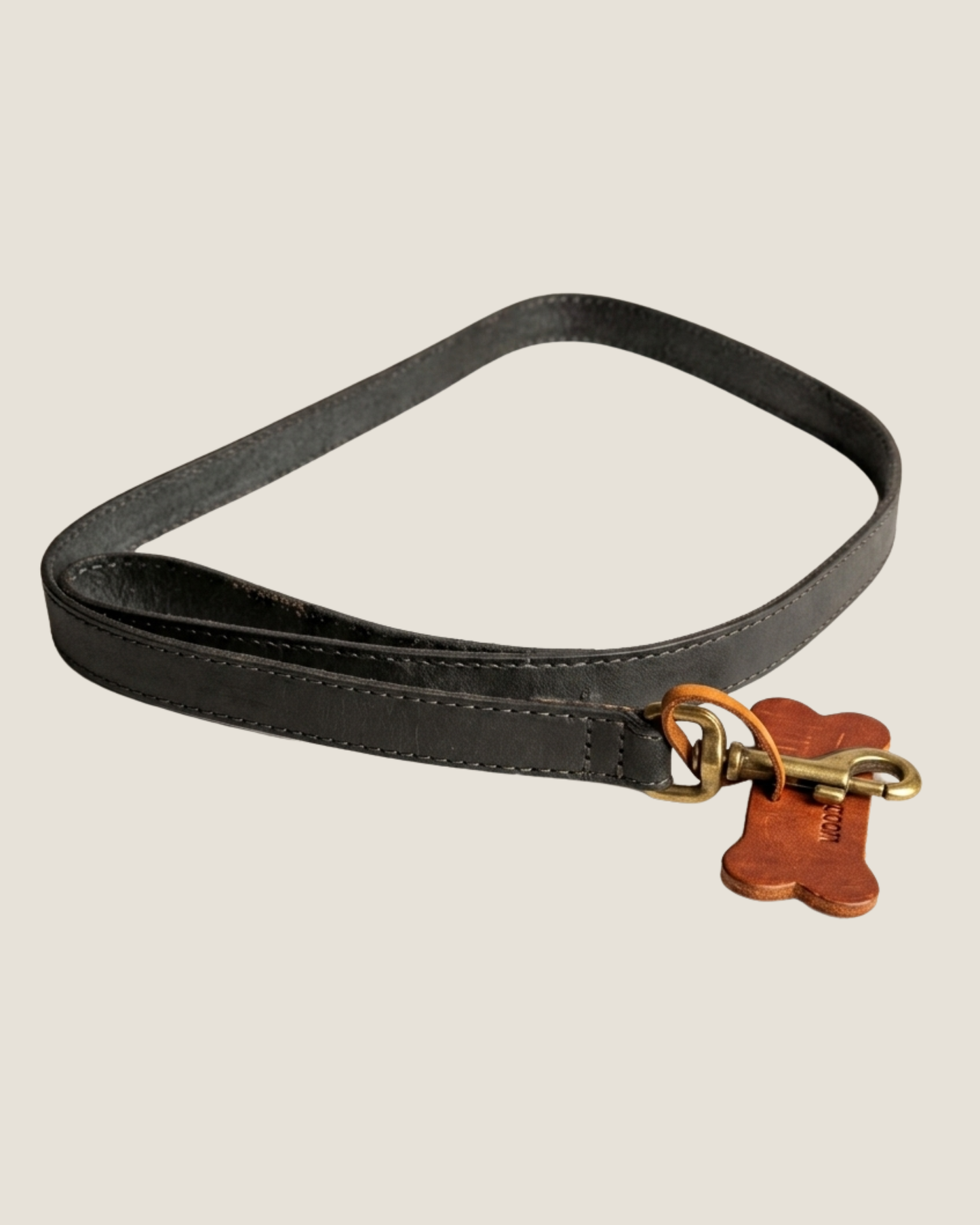 Introducing The Workroom's FLAT LEATHER DOG LEAD: a sleek black premium leather dog leash, expertly crafted with sturdy hardware and accented by a stylish brown bone-shaped tag near the metal clasp. This elegant flat leash is neatly coiled to display its refined design.