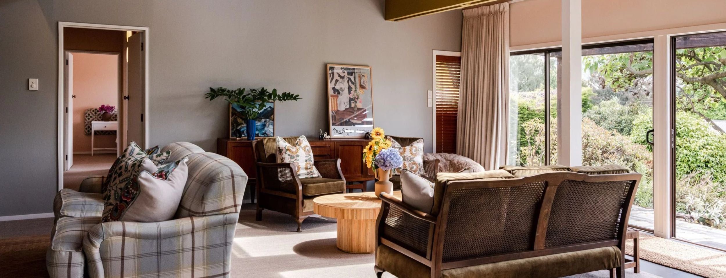 A cozy living room with vintage-style furniture, including plush armchairs and a wooden coffee table. Large windows let in natural light and overlook a garden. A painting hangs on the wall, and fresh flowers adorn the space.