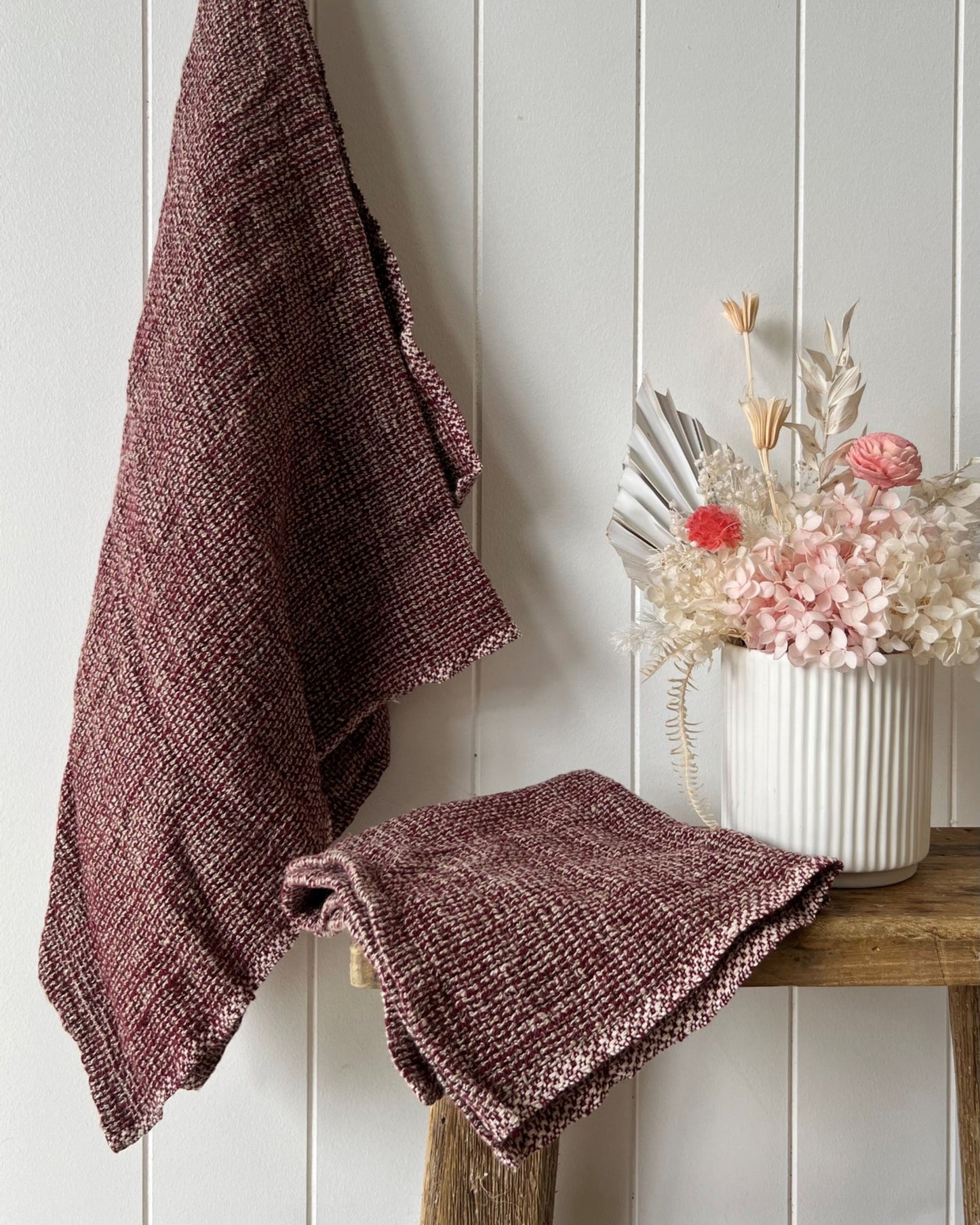 The AUDREY LINEN HAND TOWEL in a rich burgundy shade from Rustic Linen is elegantly displayed draped over a wooden stool set against a white paneled wall, enhancing the cozy, rustic ambiance. Nearby, a white vase holds dried flowers with pink blooms and pampas grass, adding to the warmth of the scene with its complementing tones.