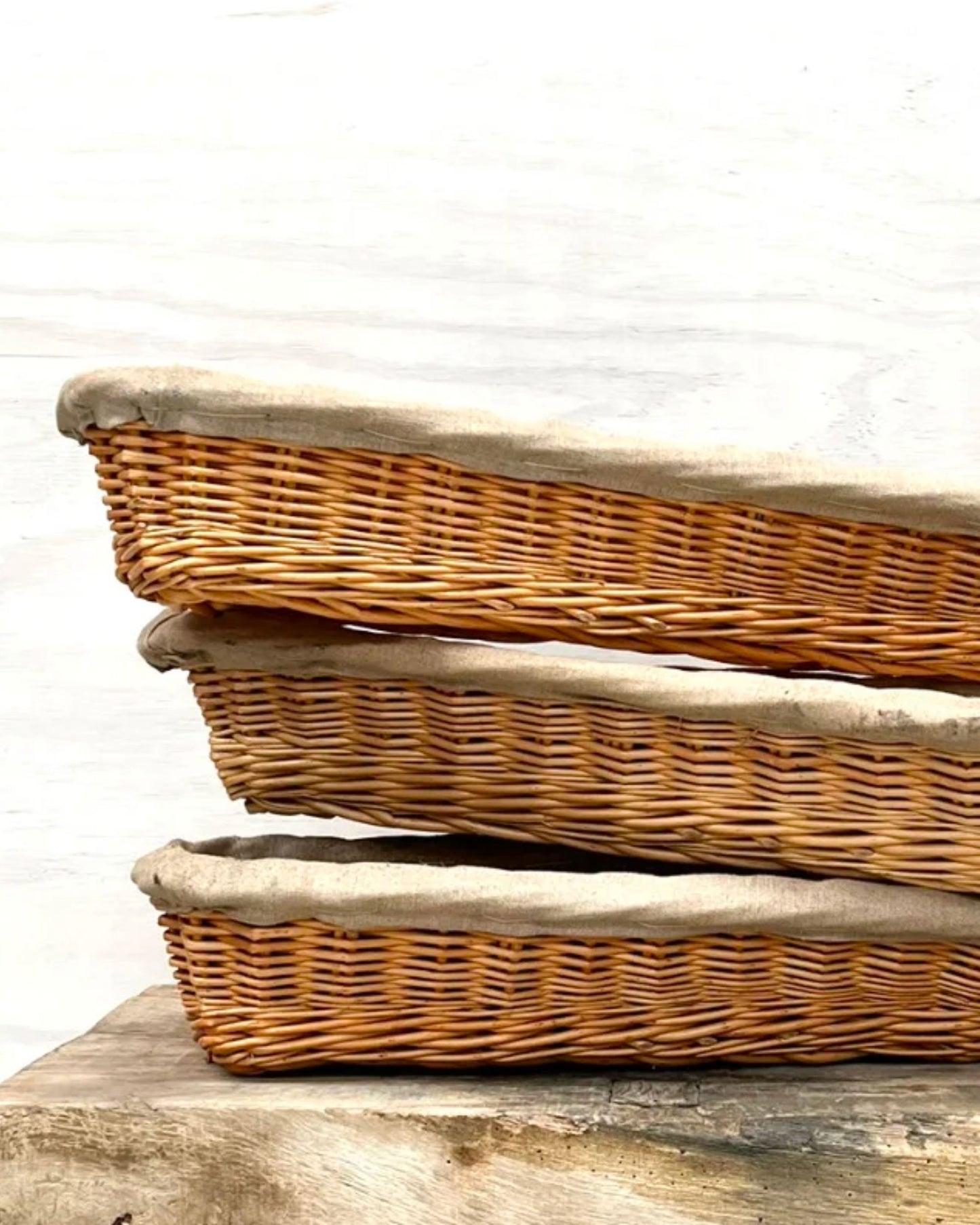 A set of three VINTAGE FRENCH BAGUETTE PROOFING BASKETS by All Things French serves as a charming decorative accent on a wooden surface. Their rustic French country style and simple elegance stand out beautifully against the plain white wall in the background.