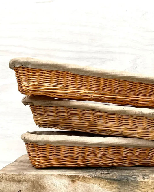 A set of three VINTAGE FRENCH BAGUETTE PROOFING BASKETS by All Things French serves as a charming decorative accent on a wooden surface. Their rustic French country style and simple elegance stand out beautifully against the plain white wall in the background.