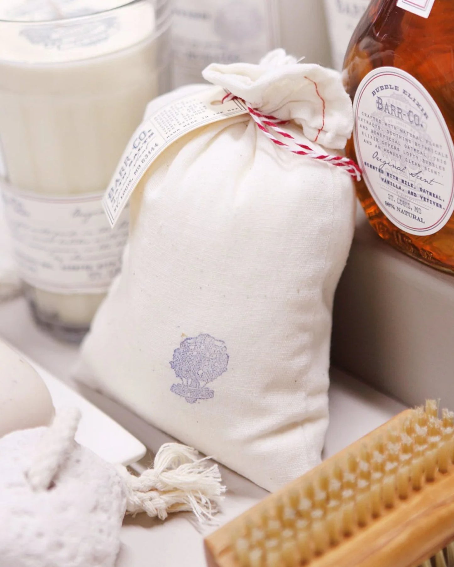 A neatly arranged bathroom scene featuring a white fabric pouch tied with string, BARR CO BATH SALTS from Barr Co, a glass candle, a bottle of bubble elixir, and a wooden brush with soft bristles. The setting exudes a clean, cozy aesthetic.