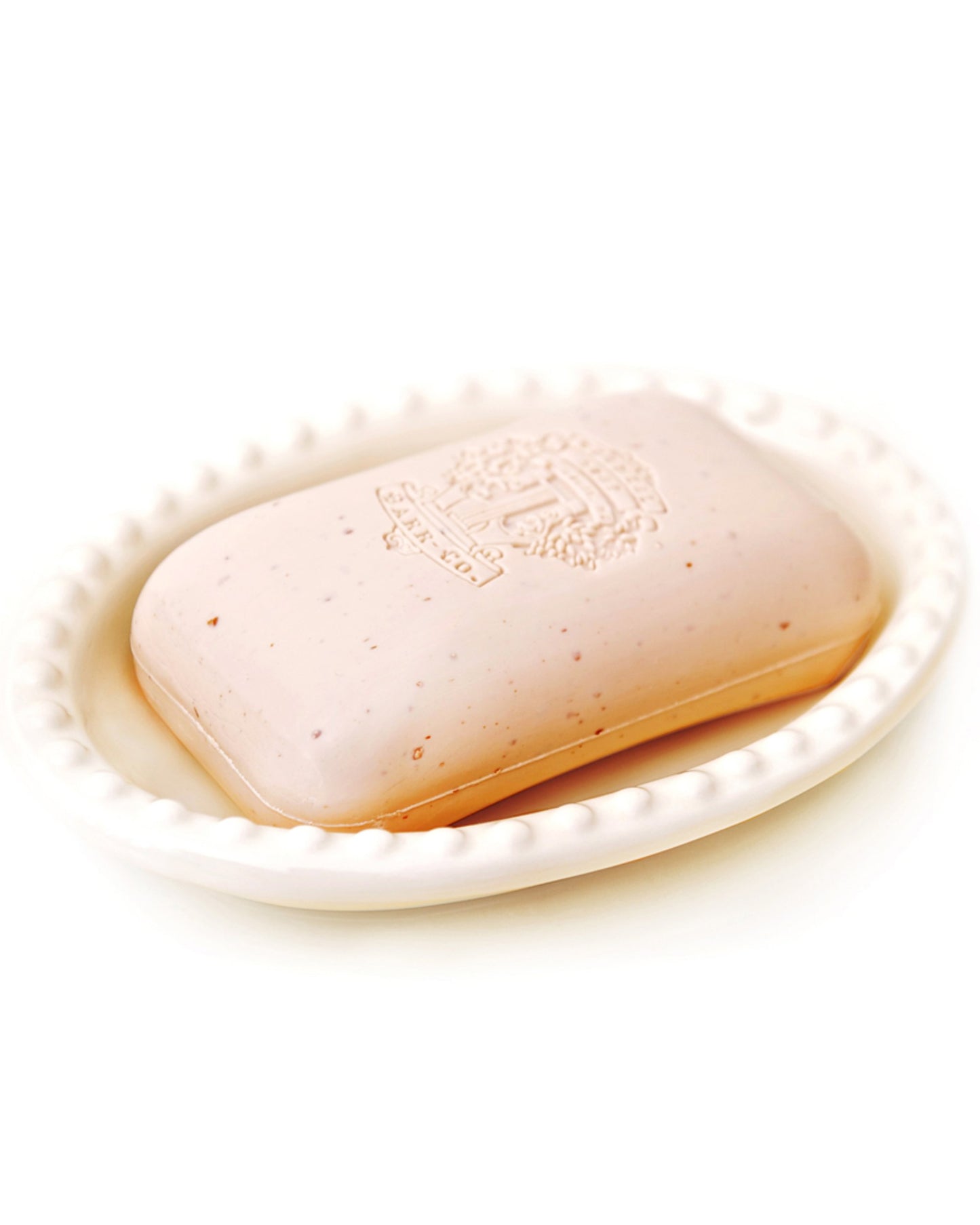A bar of Barr Co's BARR CO ORIGINAL SADDLE SOAP, embossed with text and crafted from natural vegetable ingredients, sits elegantly on a white, textured dish. Its light cream hue is accentuated by delicate speckles of oatmeal that lend a rustic charm.