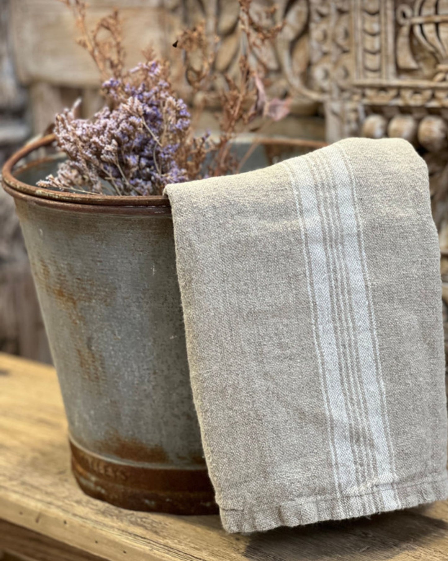 A rustic metal bucket holds dried lavender on a wooden surface. Draped over it is a BASICS WHITE STRIPE TEA TOWEL by Rustic Linen, crafted from stonewashed linen. The background showcases an ornate, textured pattern, adding charm to this delightful kitchen accessory.