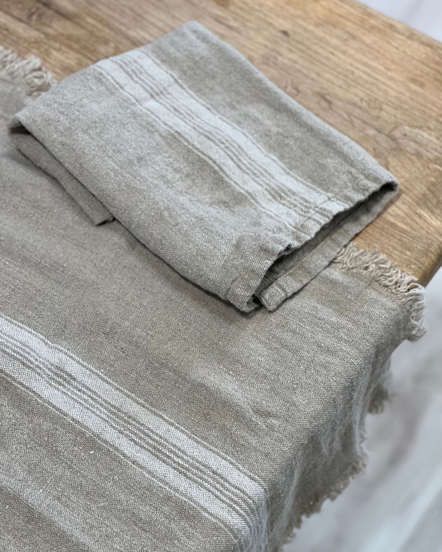 A folded gray blanket with white stripes rests on a rustic wooden surface, echoing the cozy charm of a Rustic Linen Basics White Stripe Tea Towel. Its fringed edges and textured fabric bring to mind the inviting comfort of stonewashed linen.