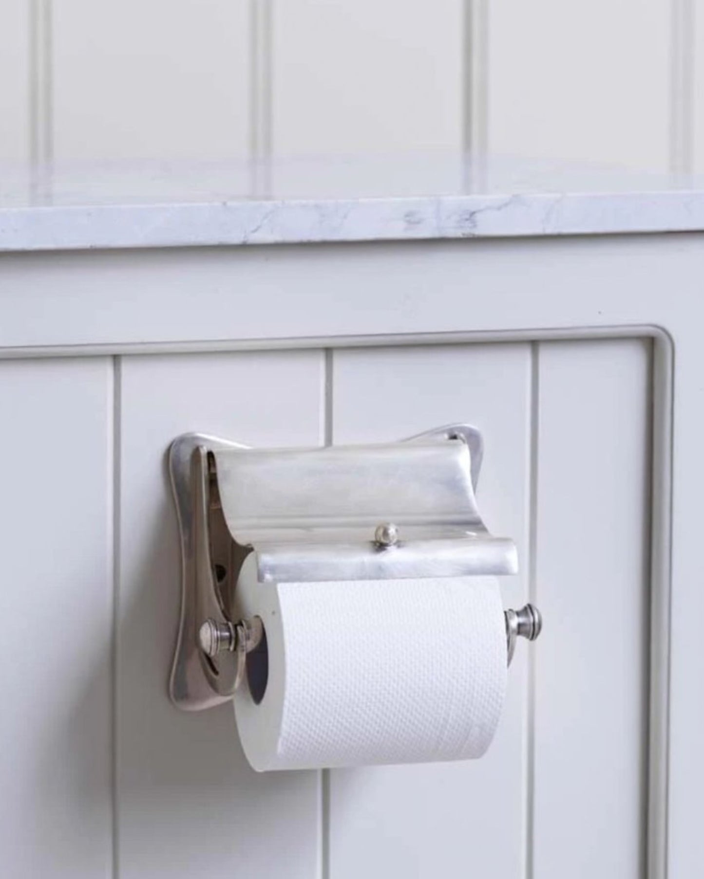A roll of toilet paper rests elegantly on the BERNARD TOILET ROLL HOLDER by Society Ink, mounted on a white paneled wall beneath a light-colored marble countertop, complemented by a sleek wall soap dispenser.