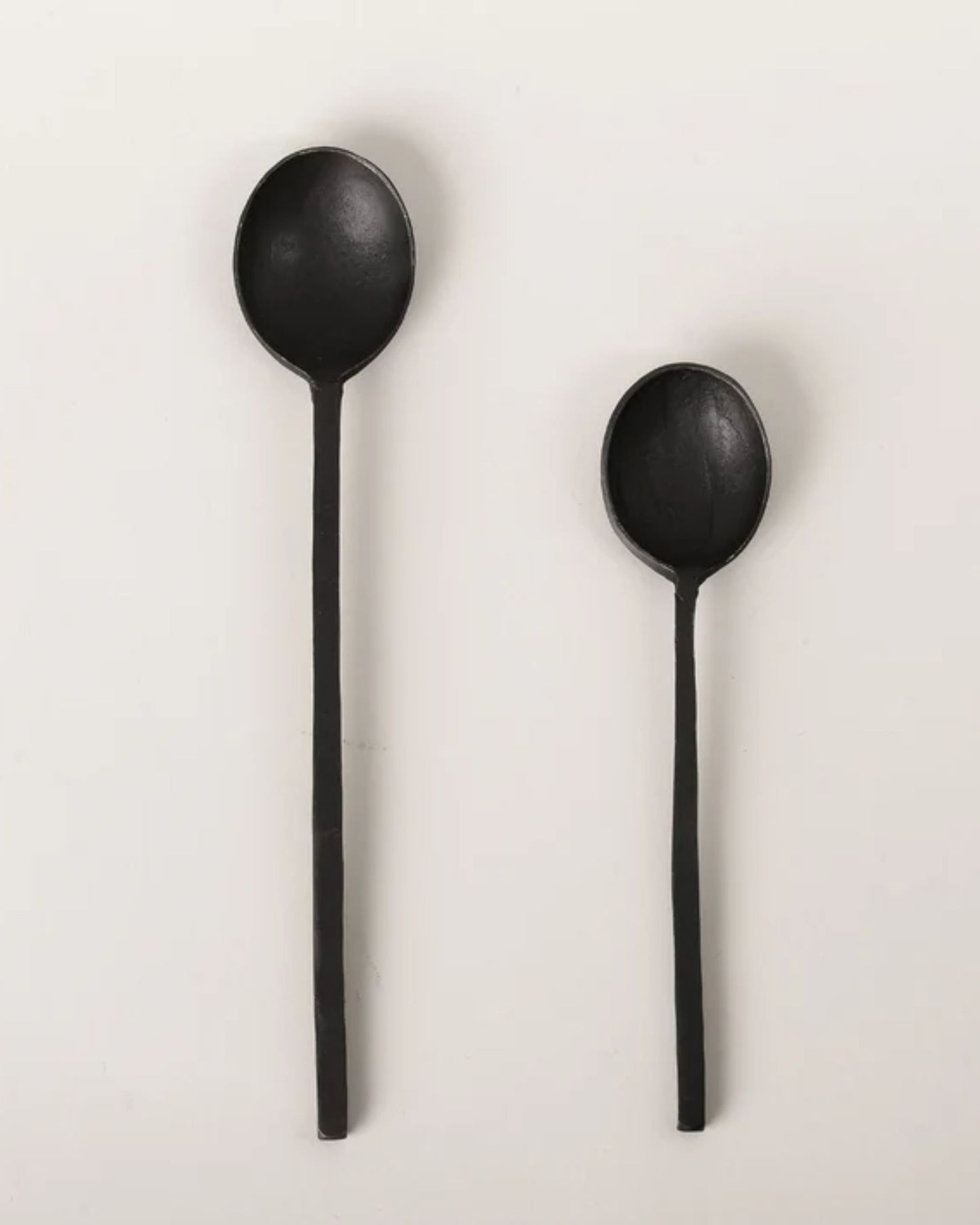 Two black spoons from New Direction are displayed on a light gray background. The one on the left is the "Black Spoon with Square Handle," featuring a larger bowl and a distinctive square handle for a unique touch, while the smaller spoon retains its elegant charm. Both showcase a simple yet sophisticated design.