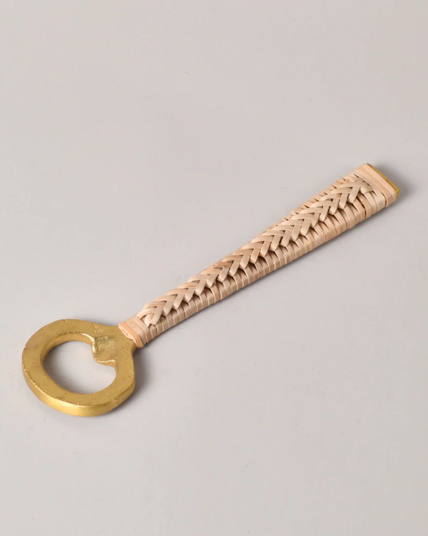The BRASS BOTTLE OPENER W/ RATTAN HANDLE by New Direction, featuring artisanal craftsmanship and a charming woven rattan handle, stands out against a neutral background, exuding rustic charm.