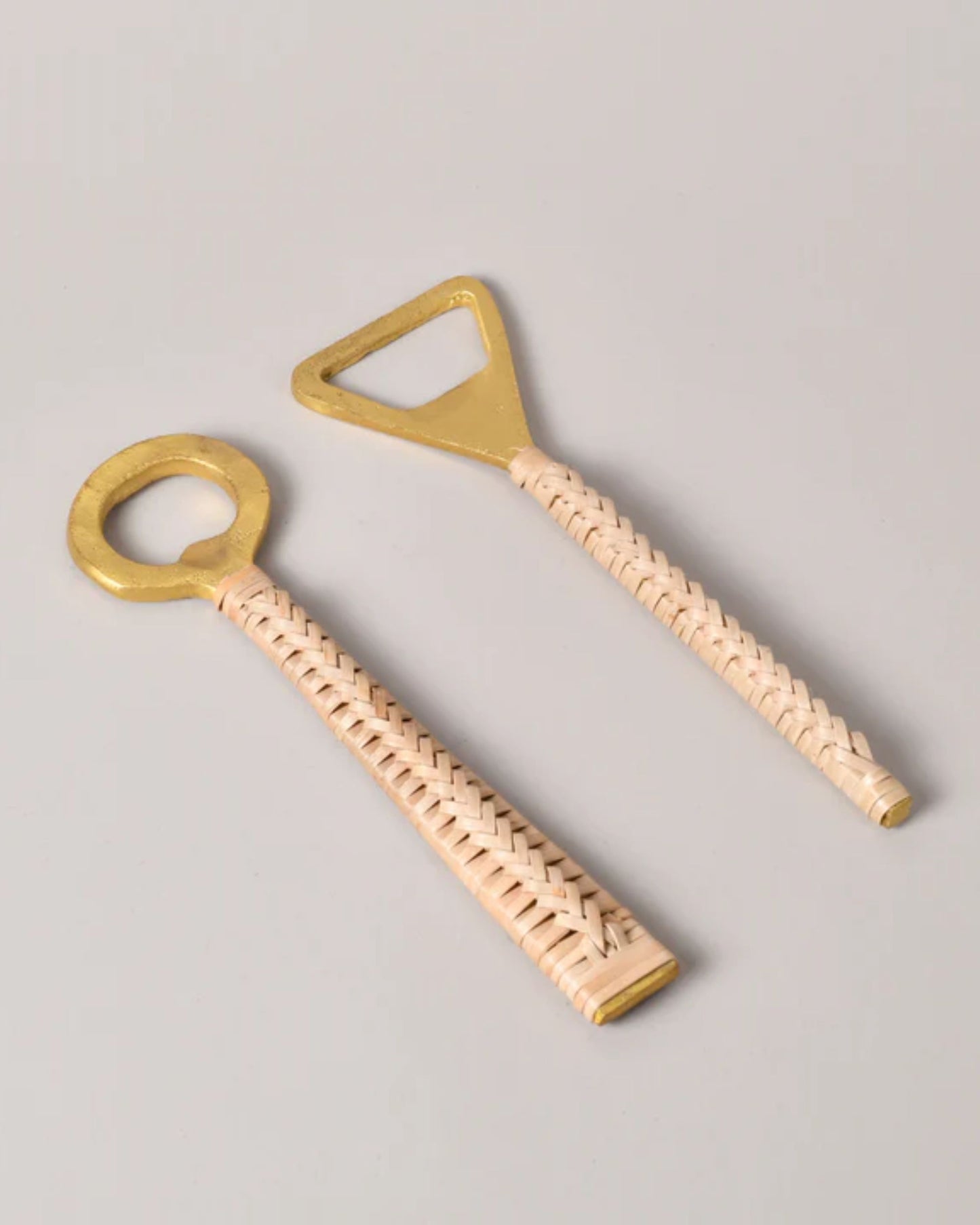 Two New Direction brass bottle openers with rattan handles exude rustic charm. The artisanal craftsmanship shines through in the Round and Triangular Brass Bottle Openers, both elegantly placed on a light gray surface.
