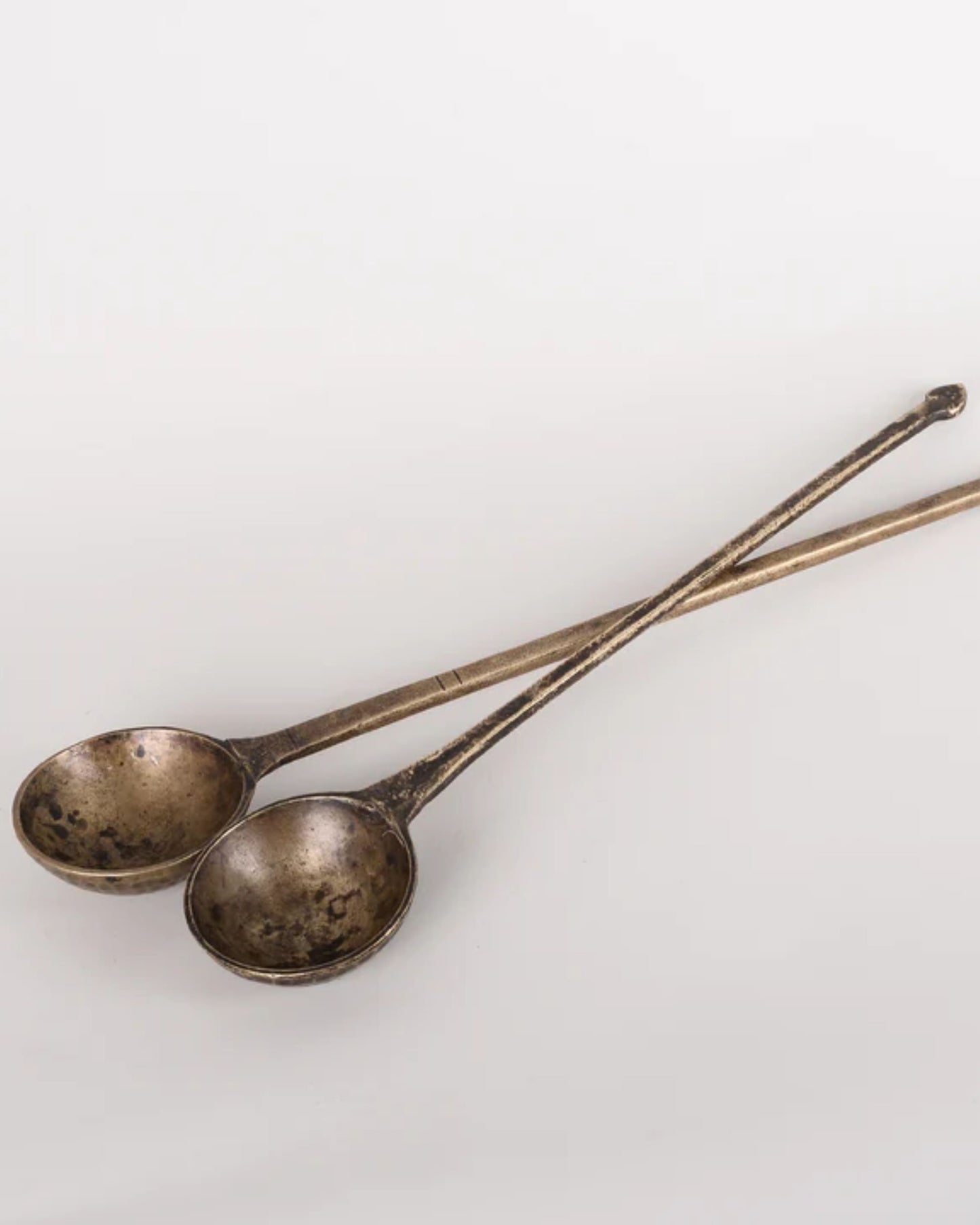 The Vintage Brass Cooking Spoons from New Direction exude antique elegance with their long handles and round, shallow bowls, artistically crisscrossed against a plain, light background.