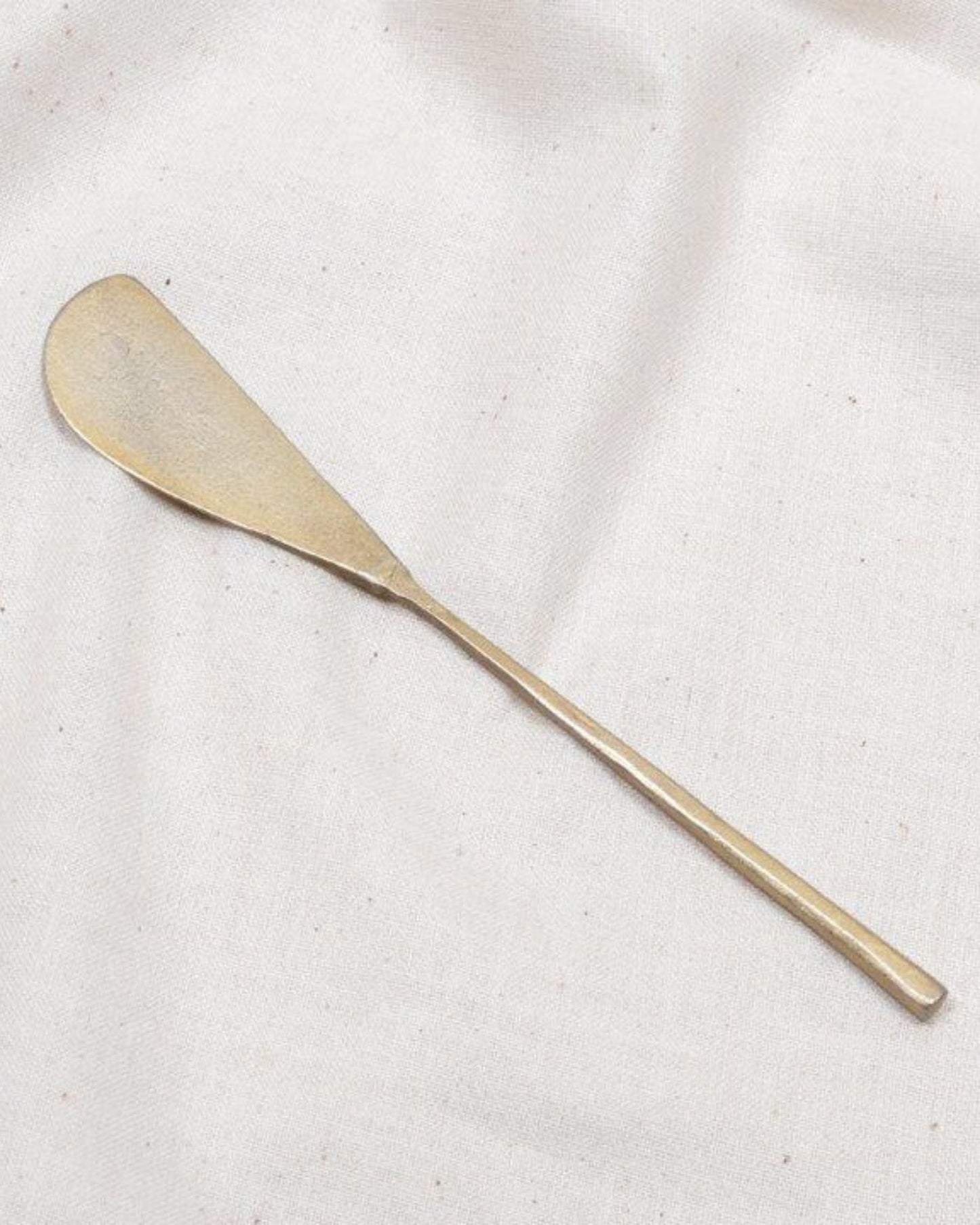 A brass shoehorn with a long, slender handle and a curved, flat end rests elegantly on a white fabric background, reminiscent of the grace of the New Direction Brass Tapas Knife amid an appetizer spread.