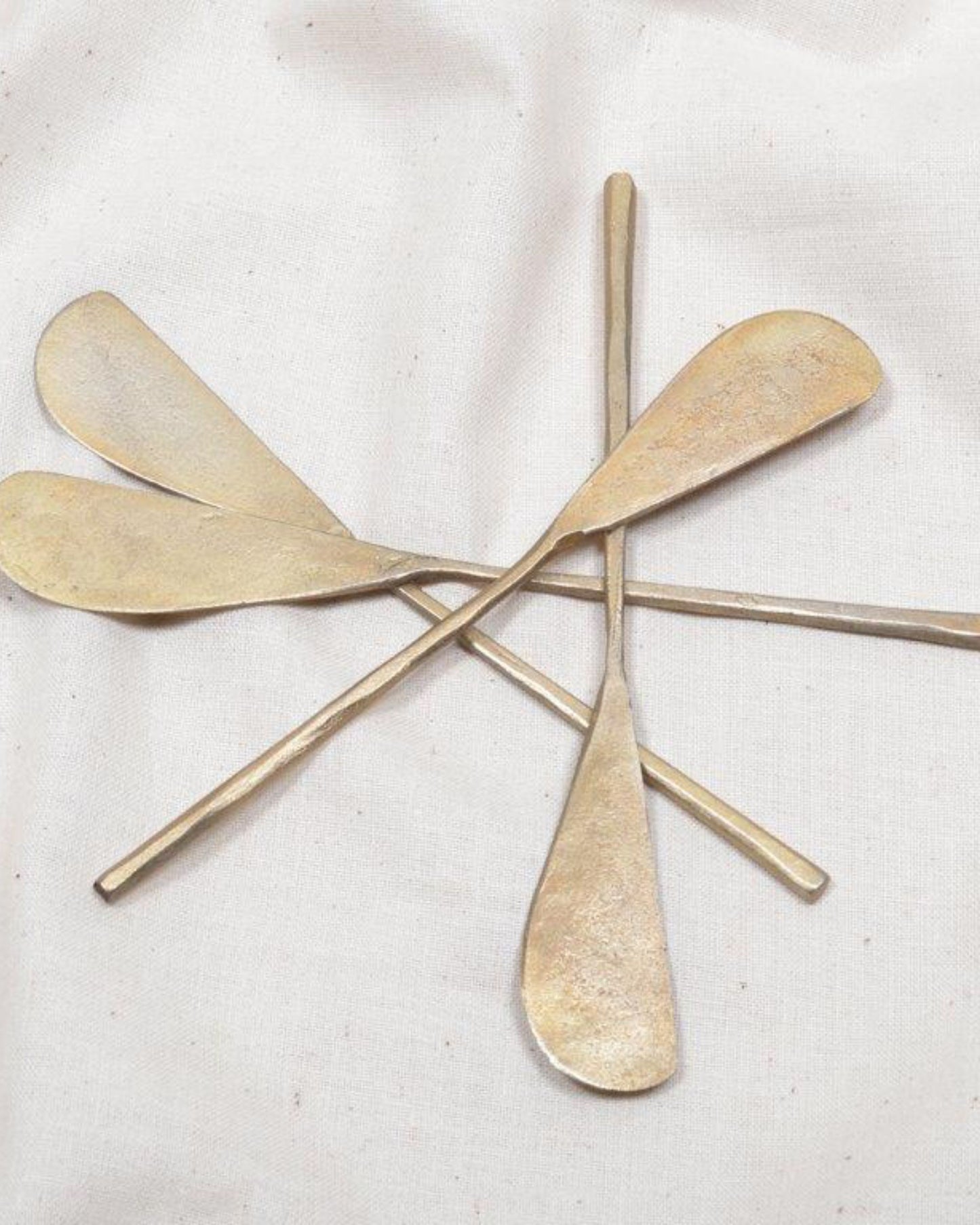 Four New Direction brass tapas knives with elongated, asymmetrical blades and straight handles are arranged in a crisscross pattern on a textured white fabric background, perfect for serving an elegant charcuterie or appetizer spread.