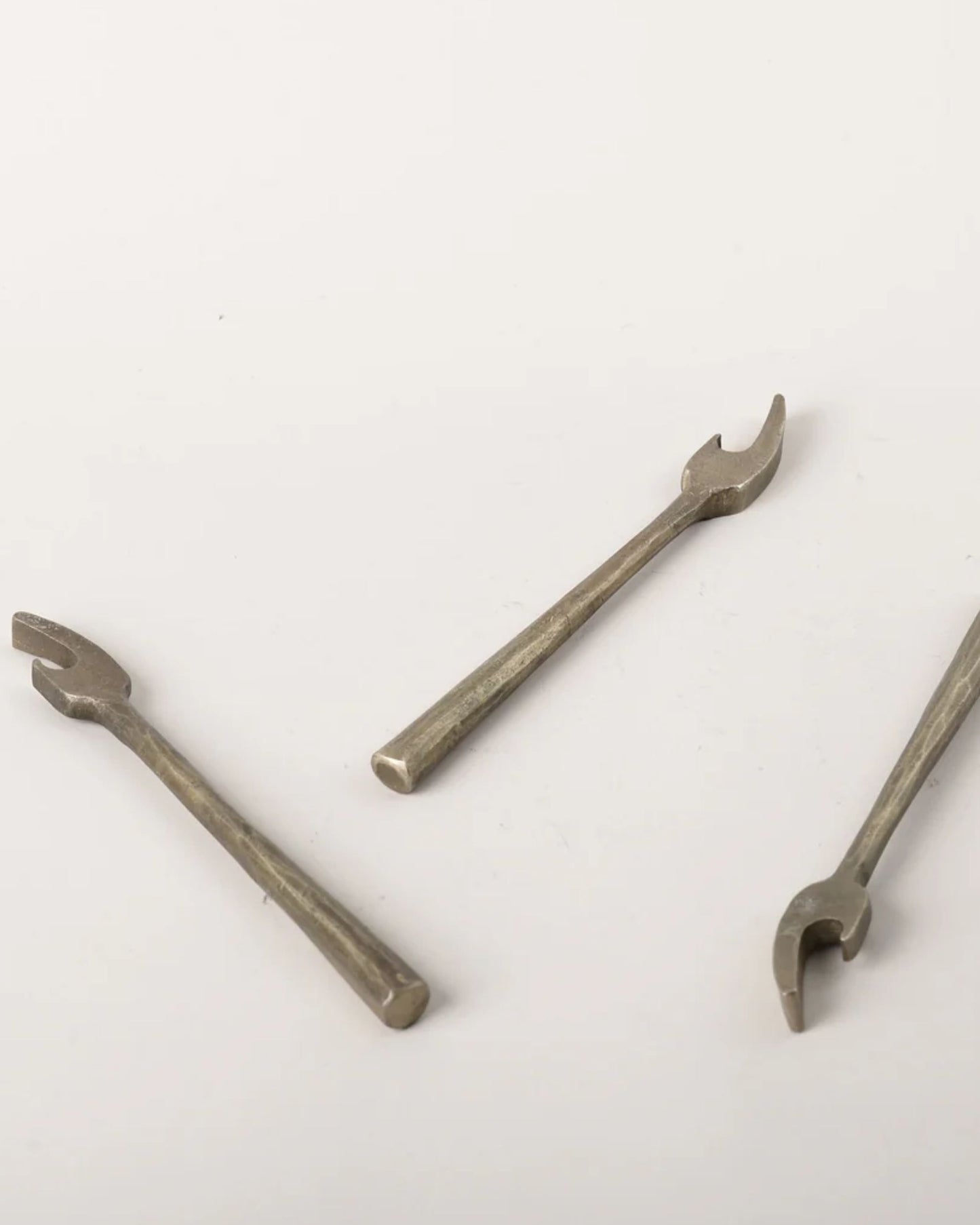 Three BRASS WRENCH BOTTLE OPENERS from New Direction, shaped like U-shaped miniature wrenches, are displayed on a plain white surface. Their textured metal surfaces exude an industrial charm with a vintage-inspired design, suggesting their potential use in machinery or craft-related tasks.
