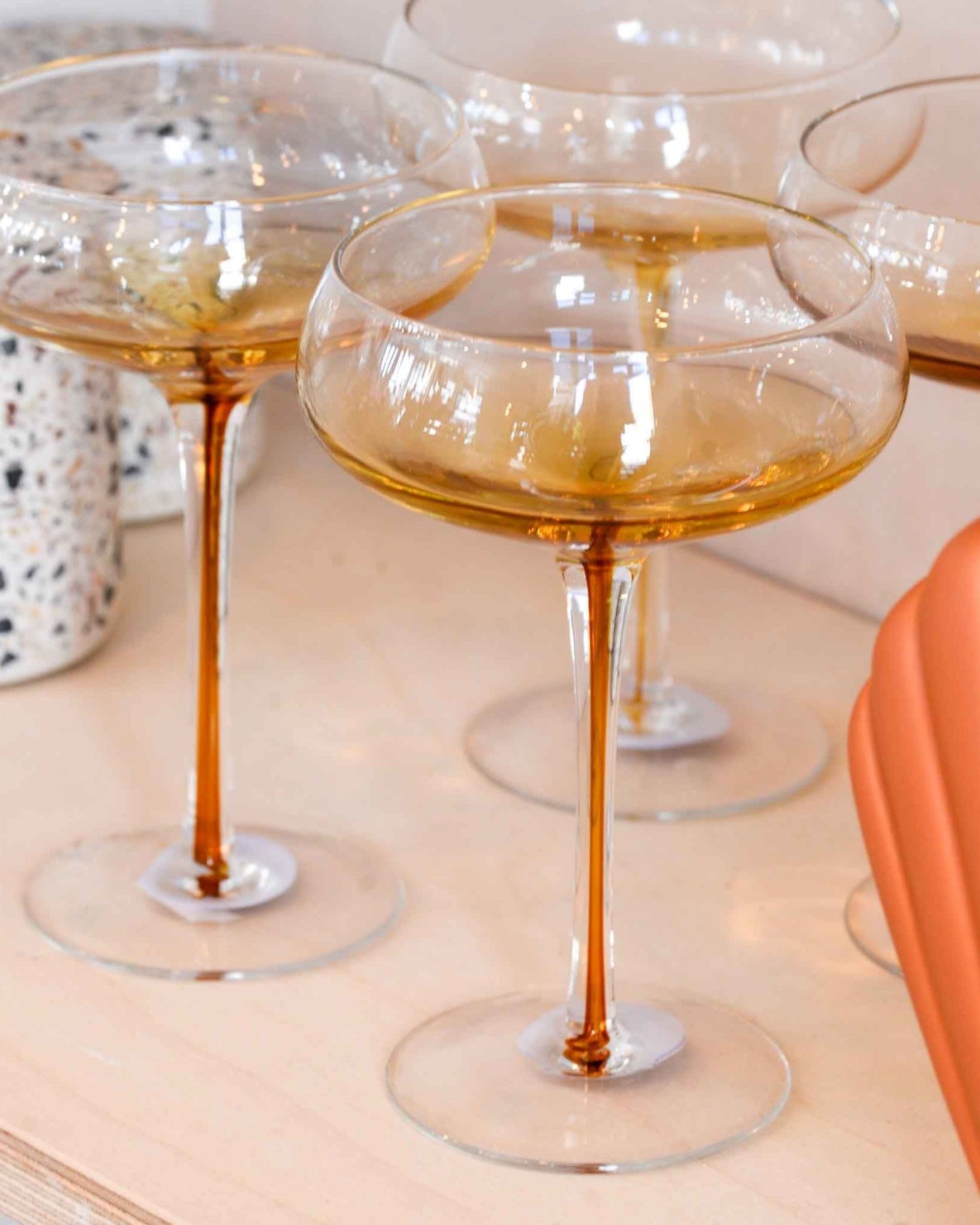 Three elegant COCKTAIL GLASSES from Broste’s collection hold amber-colored liquid, stunningly displayed on a wooden surface. These glasses, featuring long stems and round, shallow bowls, capture the essence of Nordic nature. Completing the scene in the background are a terrazzo pot and a terracotta-colored object.