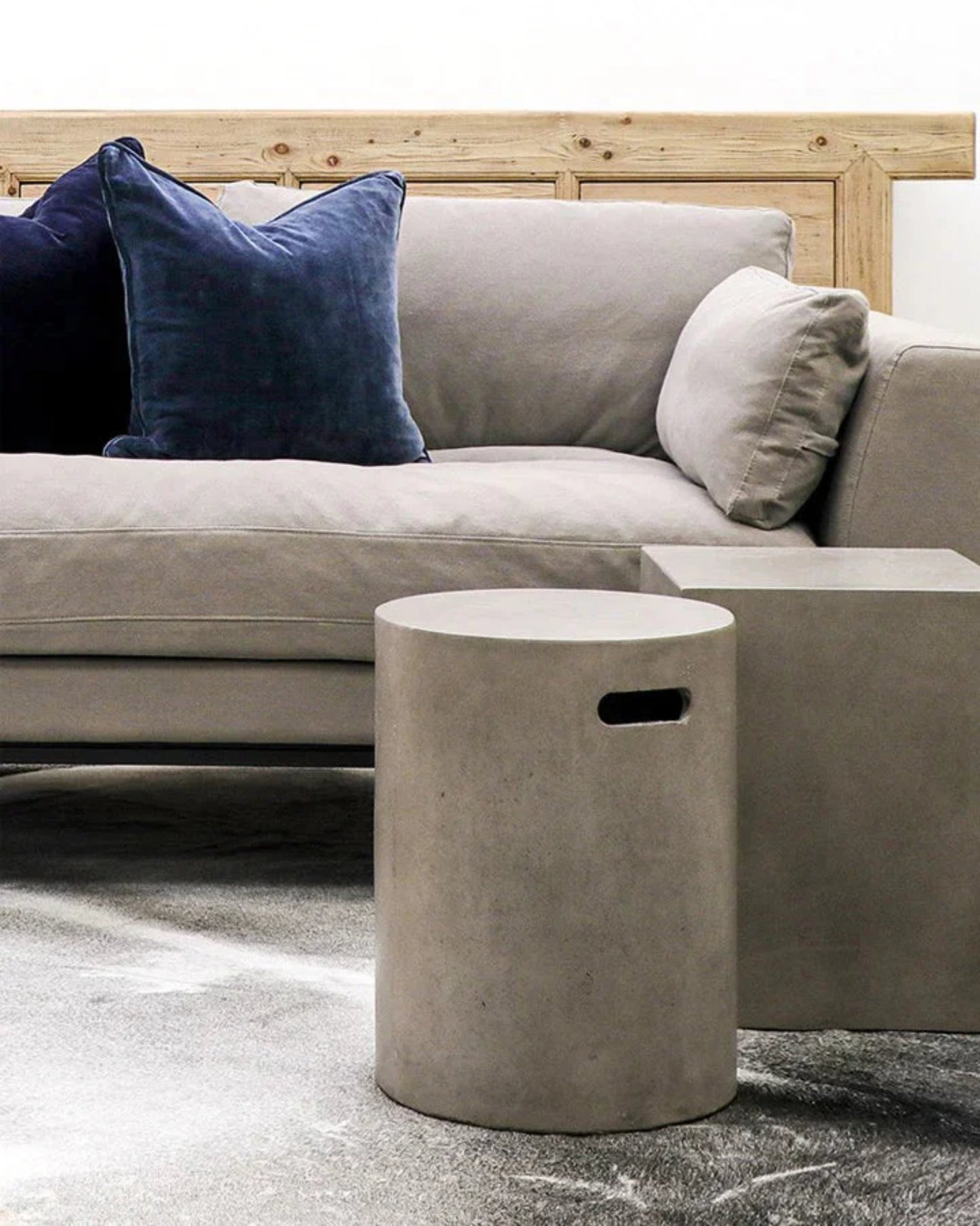 A modern living room showcases a light gray sofa adorned with dark blue and gray throw pillows. Positioned in front, two stone-colored Hawthorne CONCRETE PIPE SIDE TABLE / STOOL pieces complement the marble-like patterned floor. A versatile wooden piece can be partially seen behind the sofa.