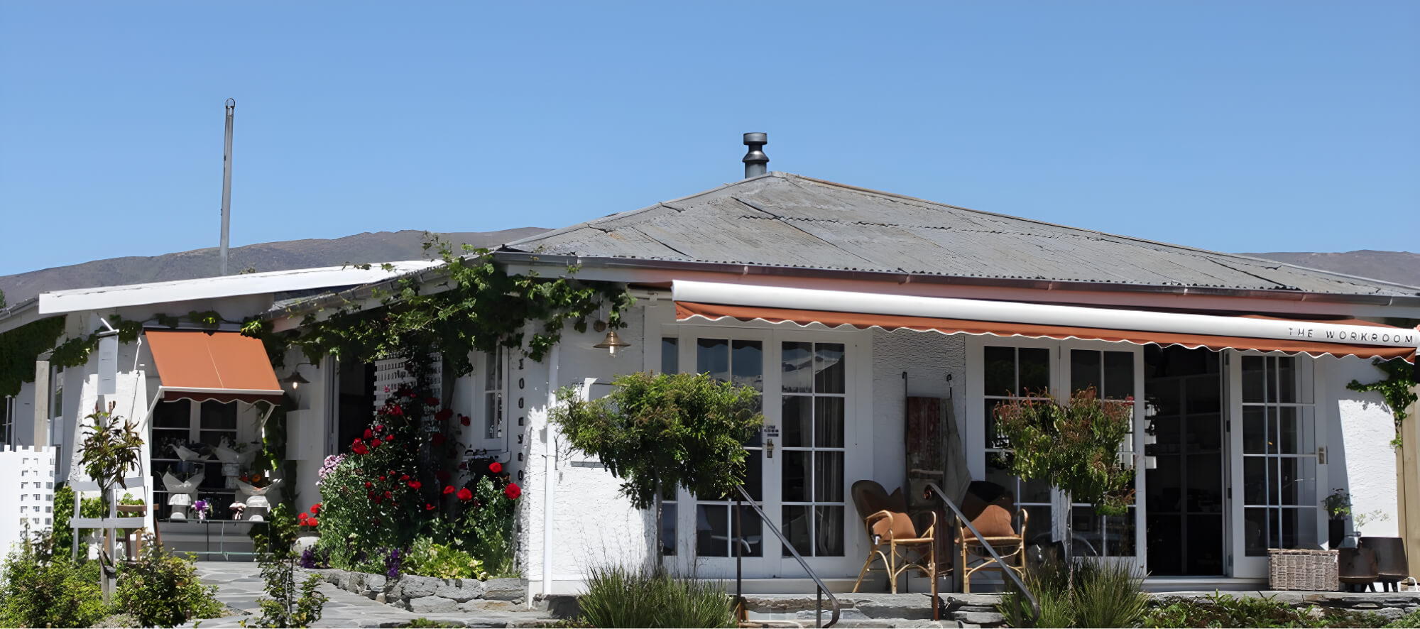 A cozy white house with a metal roof, surrounded by flowers and vines. It features large windows, a porch with chairs, and a striped awning. The sky is clear and blue in the background.