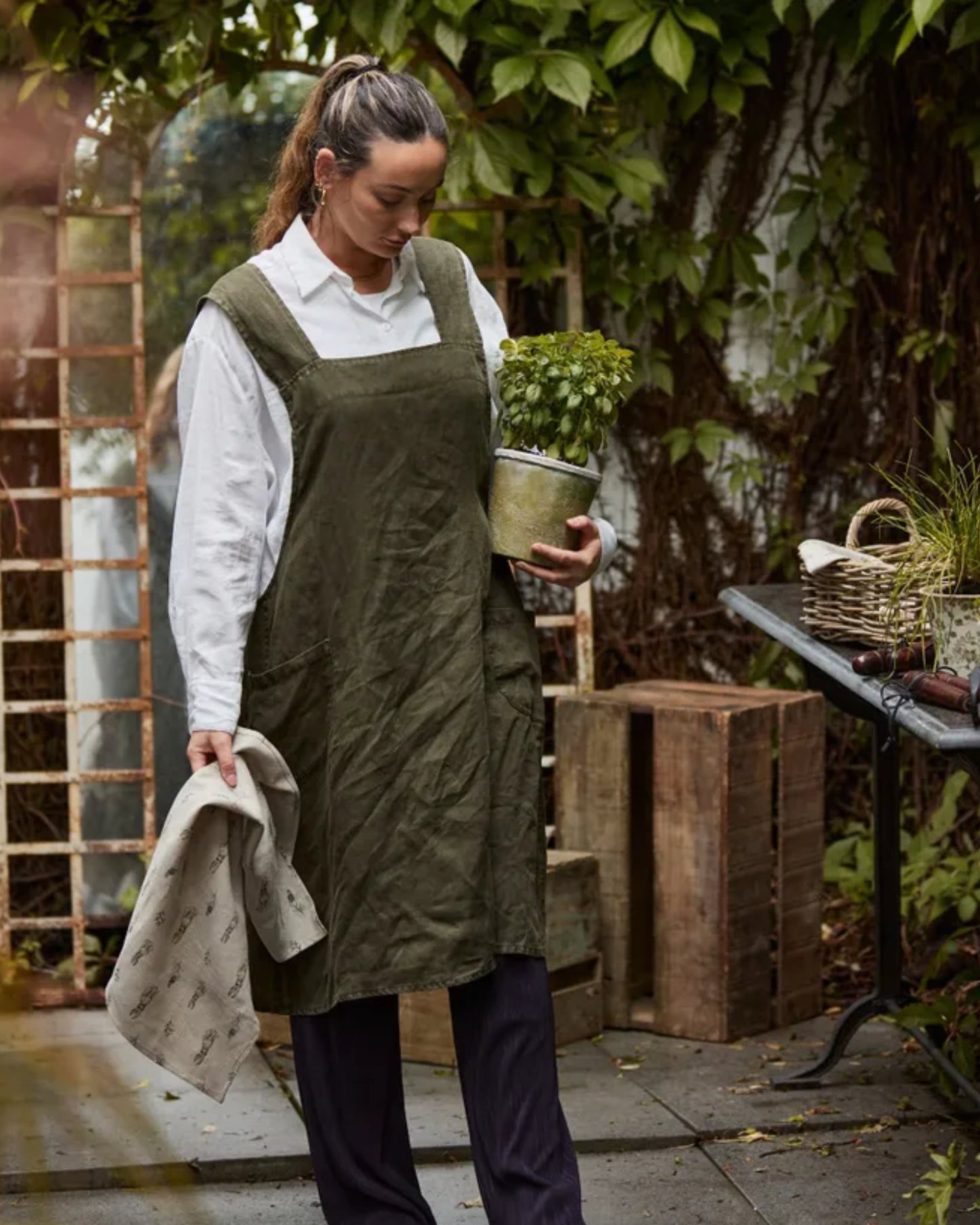 A person wearing a Cross Back Apron Stonewashed Linen by French Country holds a potted plant while standing in a garden. They are gazing down at the plant, surrounded by lush greenery, stonewashed black wooden crates, and a table with gardening tools.