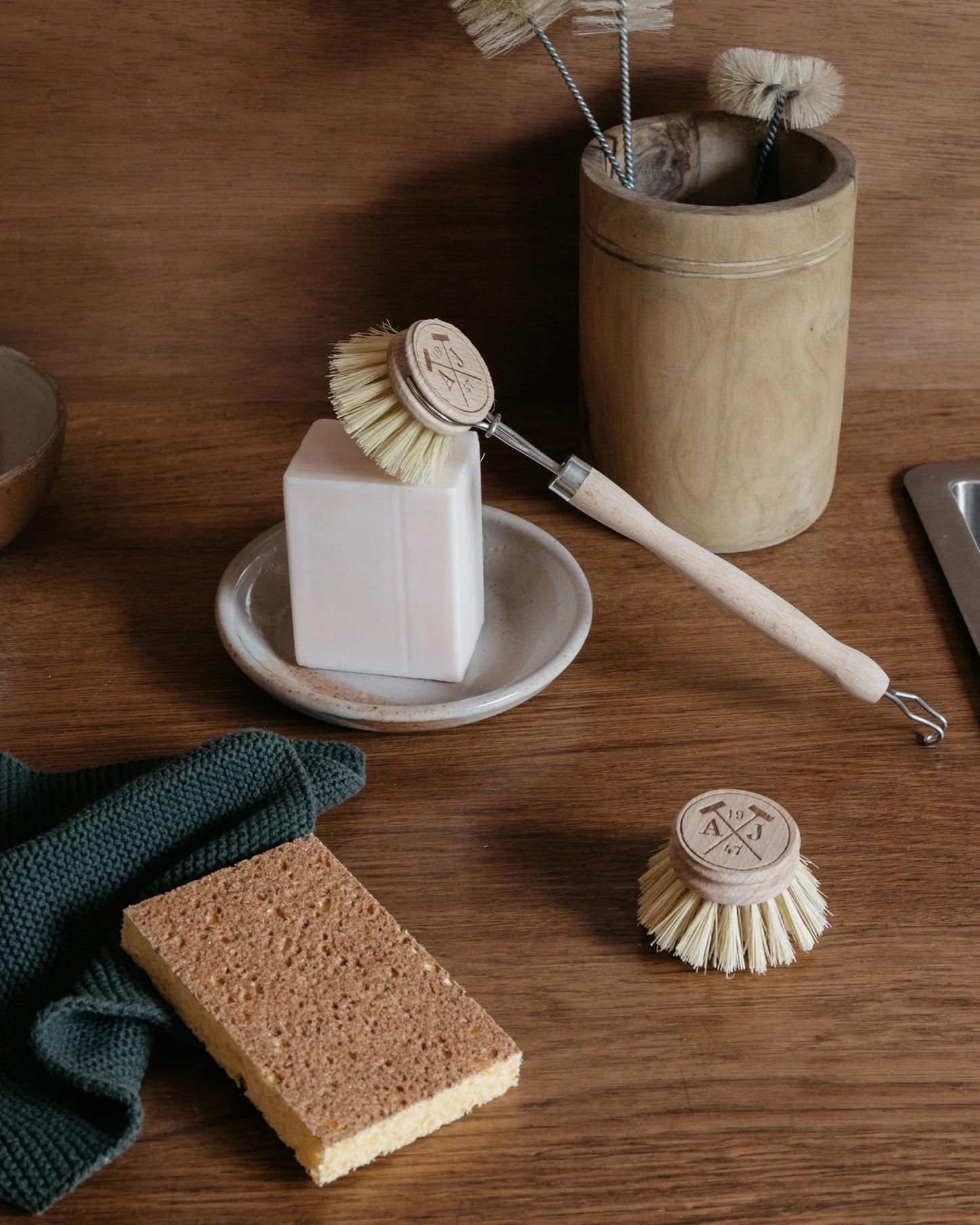 In a kitchen scene, the DISHWASHING GIFT SET by Andree Jardin is displayed, featuring a sustainable dishwashing set that includes a wooden dish brush with a detachable head and a block of soap resting on a ceramic dish. Accompanying it are a brown sponge and green cloth, while reusable items such as extra brush heads are stored neatly in a wooden container.