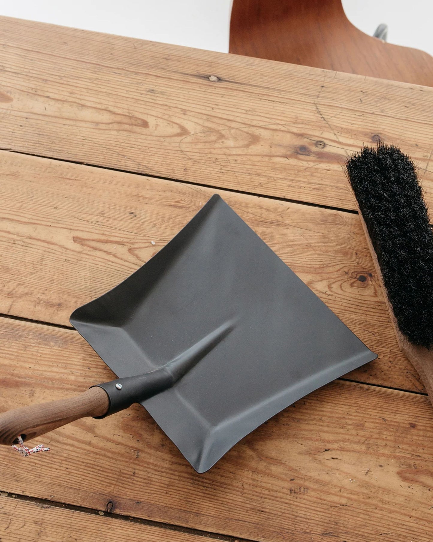 A DUSTPAN in black sheet metal, matched with an Andree Jardin natural-bristle brush boasting a French beechwood handle, sits gracefully on a wooden table. The table's minor scratches and natural finish enhance the rustic charm of this elegantly crafted cleaning set.