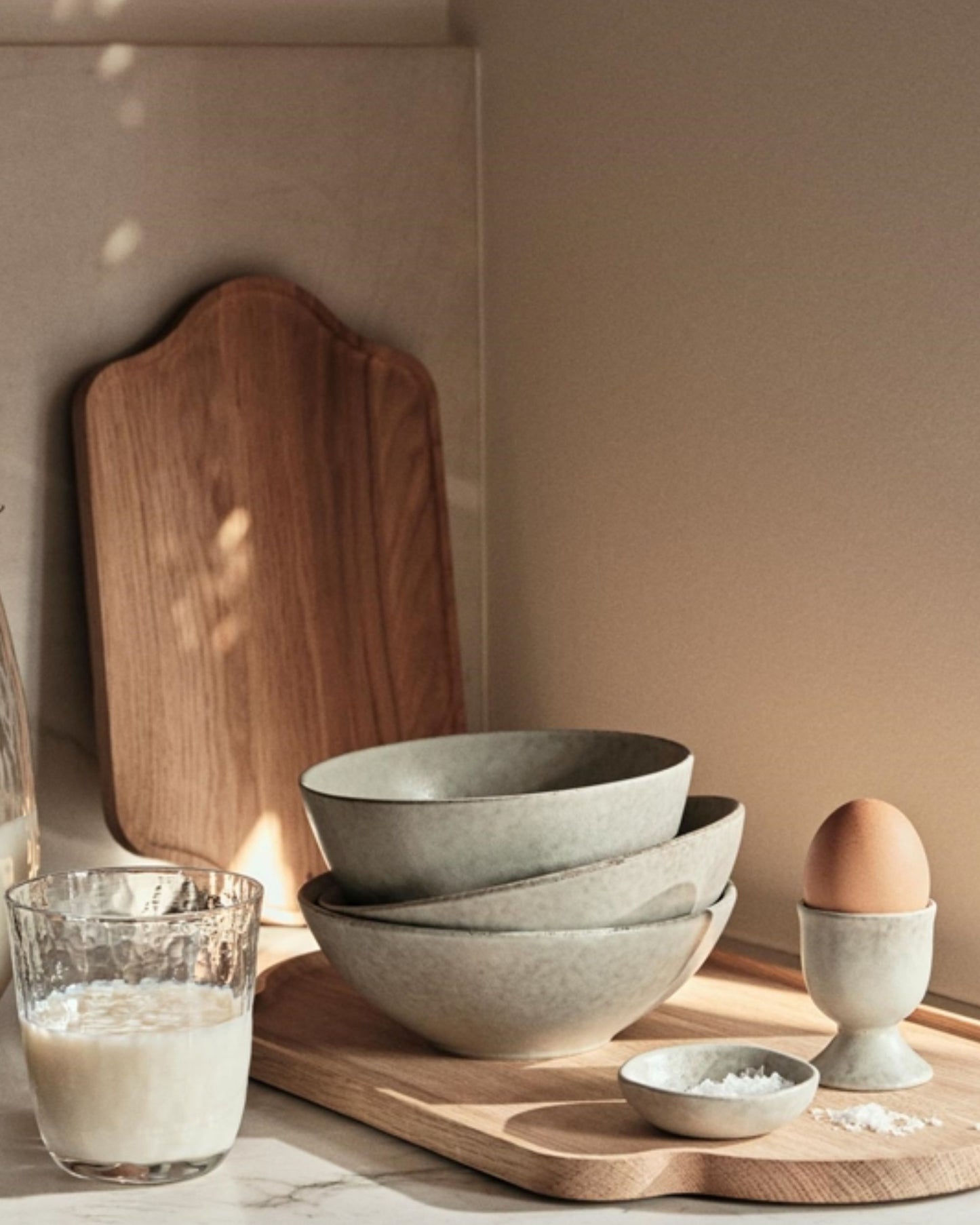 A cozy kitchen scene features ceramic bowls stacked neatly, a Broste EGG CUP holding an egg, a small dish of salt, and a glass of milk on a wooden board. A wooden cutting board leans against the wall in warm, natural lighting, creating the perfect breakfast table setting.