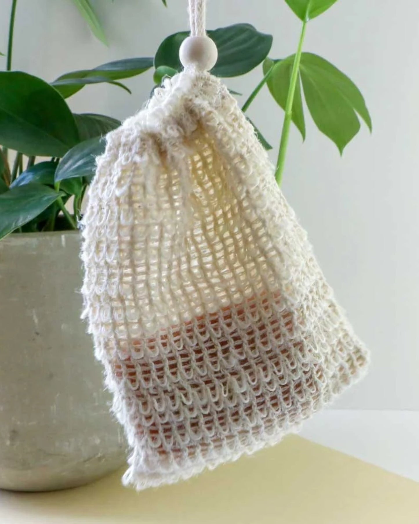 A white EXFOLIATING SOAP SACK by Redecker hangs in front of a potted plant with large green leaves. Inside the transparent knit drawstring sack, there appears to be a brown object, possibly soap, ideal for exfoliation. The background is light and neutral.