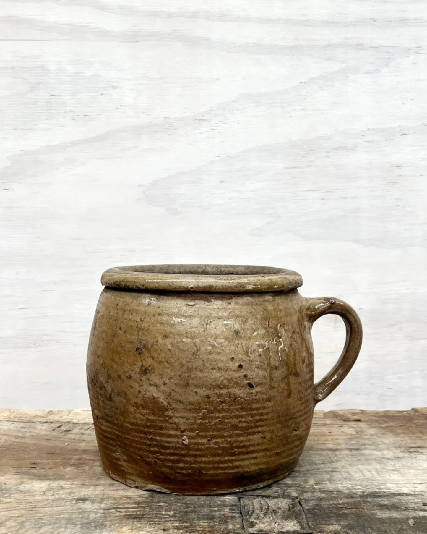 A FRENCH VINTAGE EARTHENWARE PRESERVING POT by All Things French, featuring a rustic brown ceramic design with a handle, is placed on a textured wooden surface. Its weathered appearance exudes the vintage charm of South West France, reminiscent of traditional rustic earthenware.