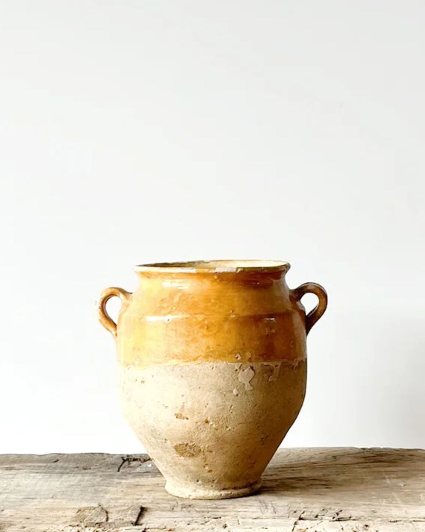 The FRENCH VINTAGE MUSTARD YELLOW CONFIT POT by All Things French showcases a rustic charm with its two handles and is glazed in a faded mustard yellow on the upper half, complemented by an unglazed, textured surface on the lower half. This piece makes an ideal centerpiece as it rests atop a weathered wooden surface against a simple white background.
