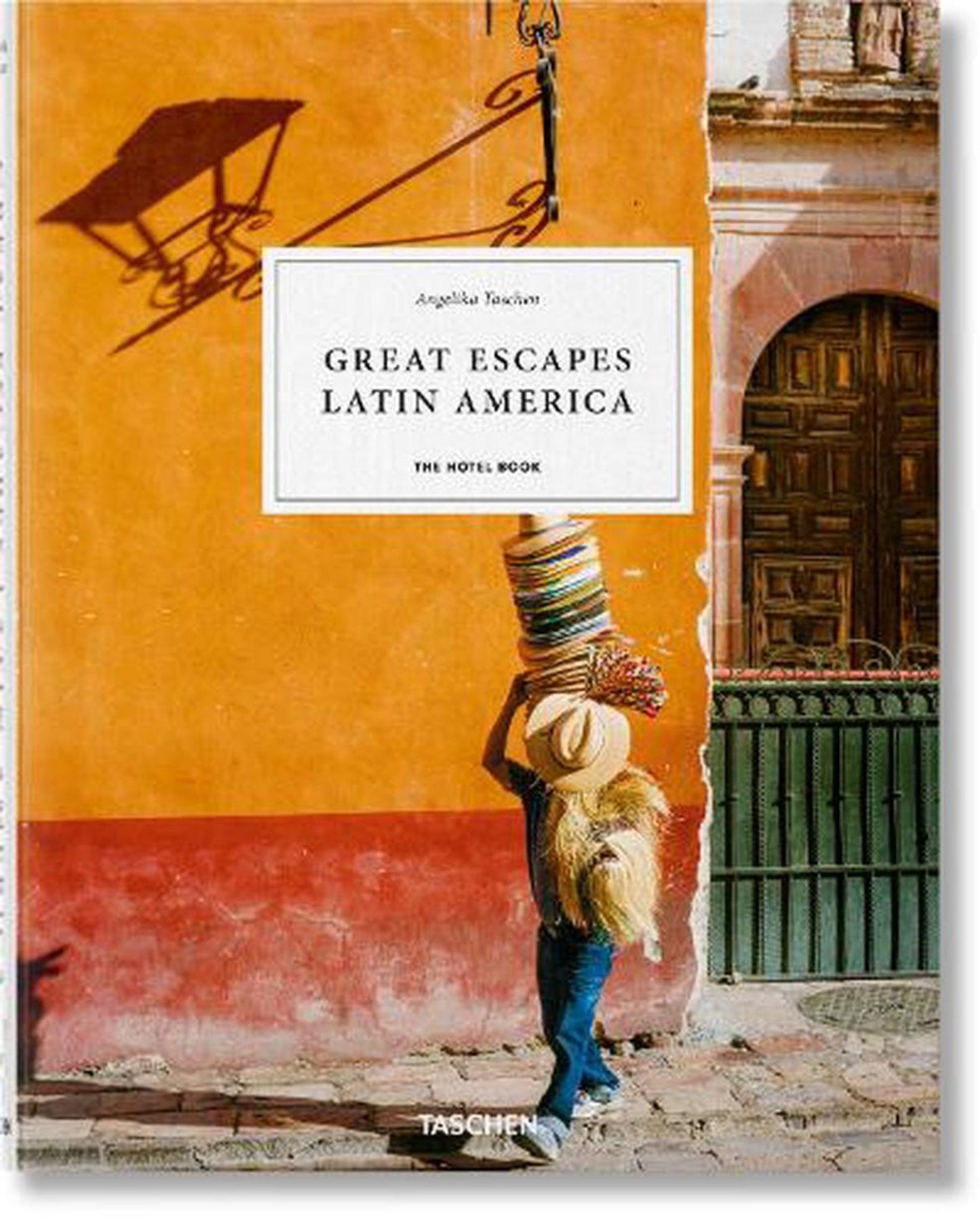 A person in jeans and a straw hat walks past an orange and red wall adorned with a decorative door, carrying colorful baskets on their head. Capturing the vibrant and multicultural essence of Latin America, the book cover is titled GREAT ESCAPES - LATIN AMERICA by Nation Wide Books.