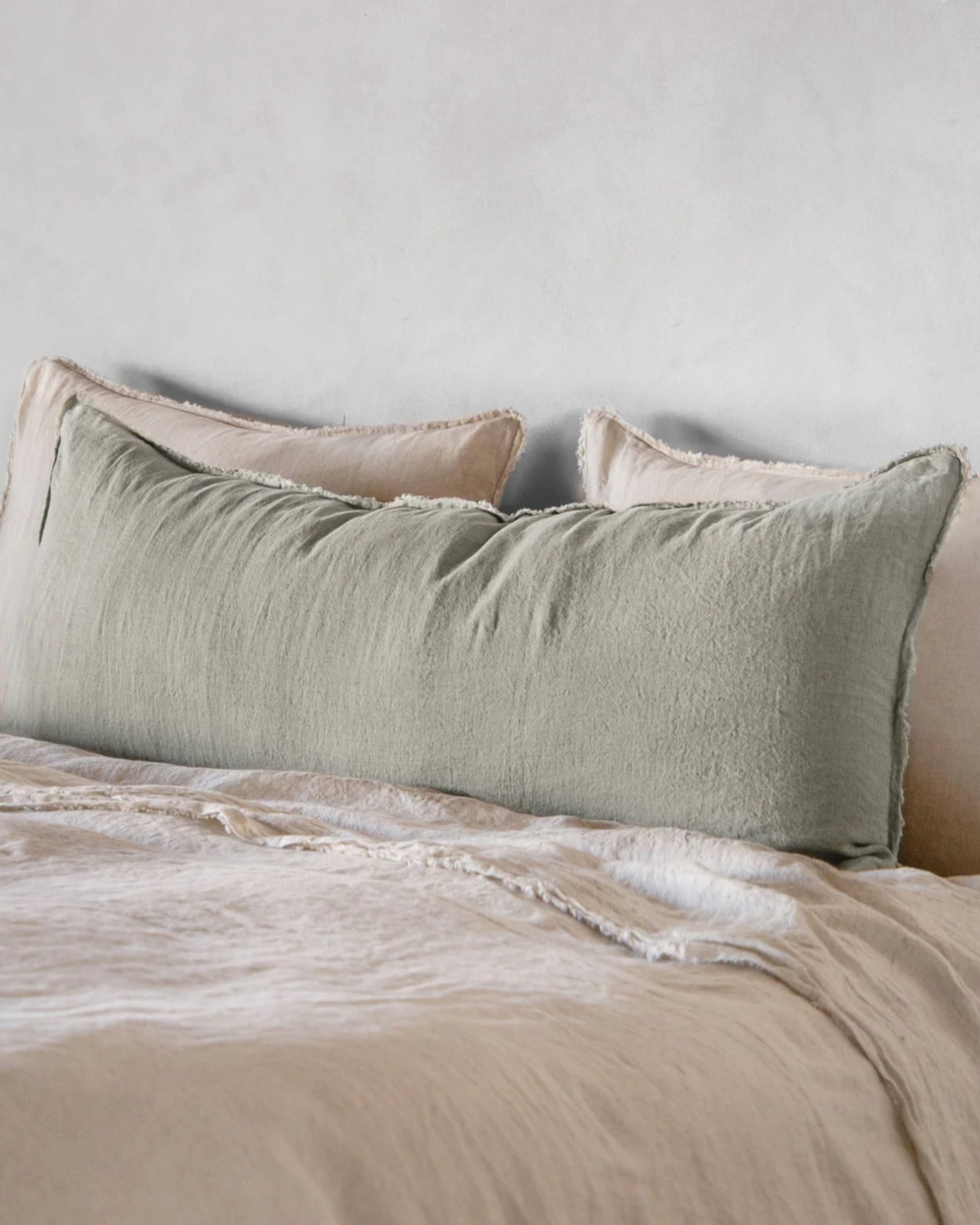 A neatly made bed features a Hale Mercantile Co's Hale Flocca Body Pillow Case in light green, placed against two beige pillows. The bed is dressed with a beige sheet and a light throw blanket, complemented by a light gray wall backdrop.