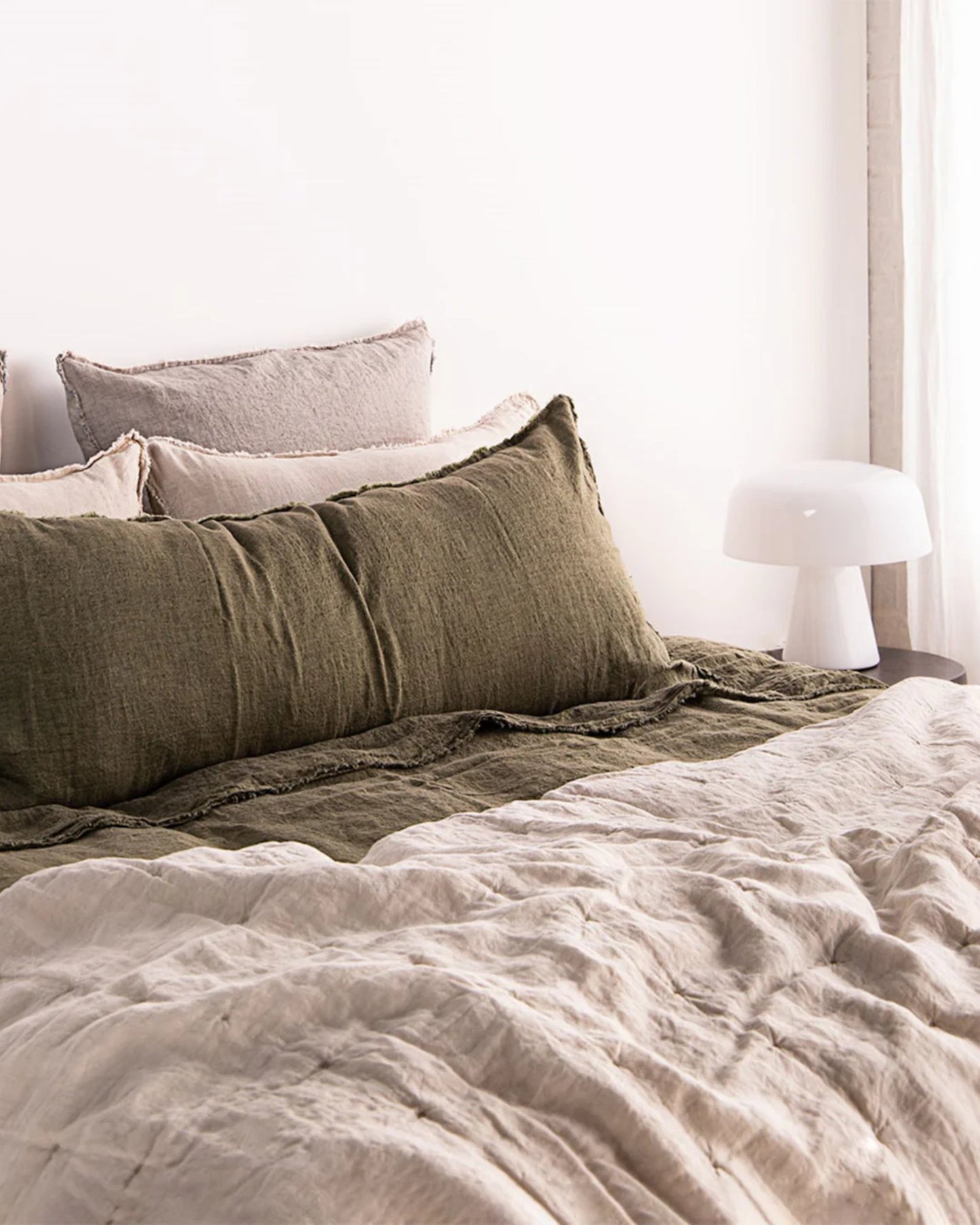 A cozy bedroom scene showcases a neatly made bed adorned with green and beige linens and pillows, including the Hale Flocca Body Pillow Case by Hale Mercantile Co. A small white lamp rests on a bedside table, set against a light-colored wall, creating a serene and inviting atmosphere.