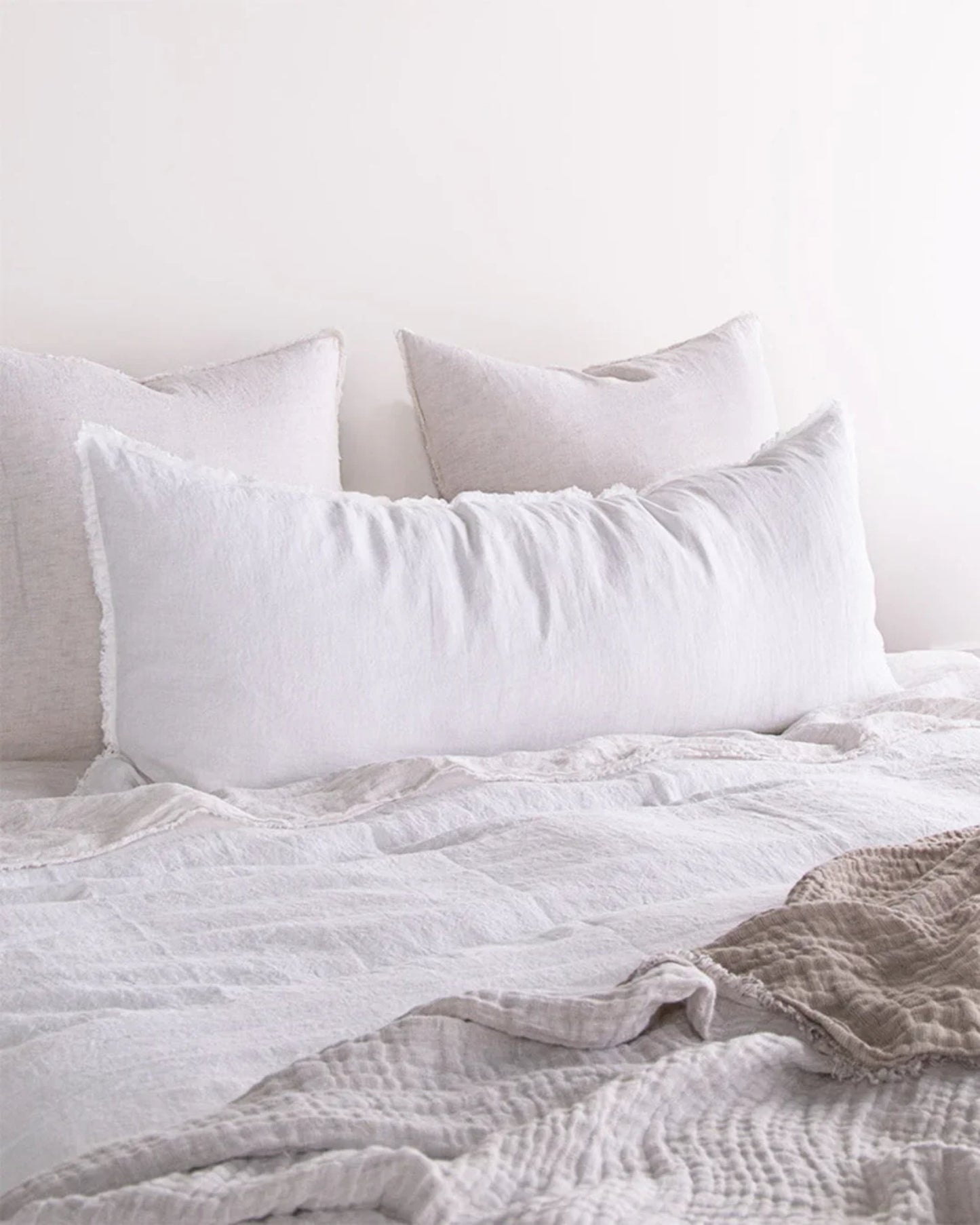 A cozy bed featuring soft, off-white bedding and the Hale Flocca Body Pillow Case in white at the head. It is accompanied by two square pillows positioned behind it, and a light beige throw is partially draped across the foot of the bed.