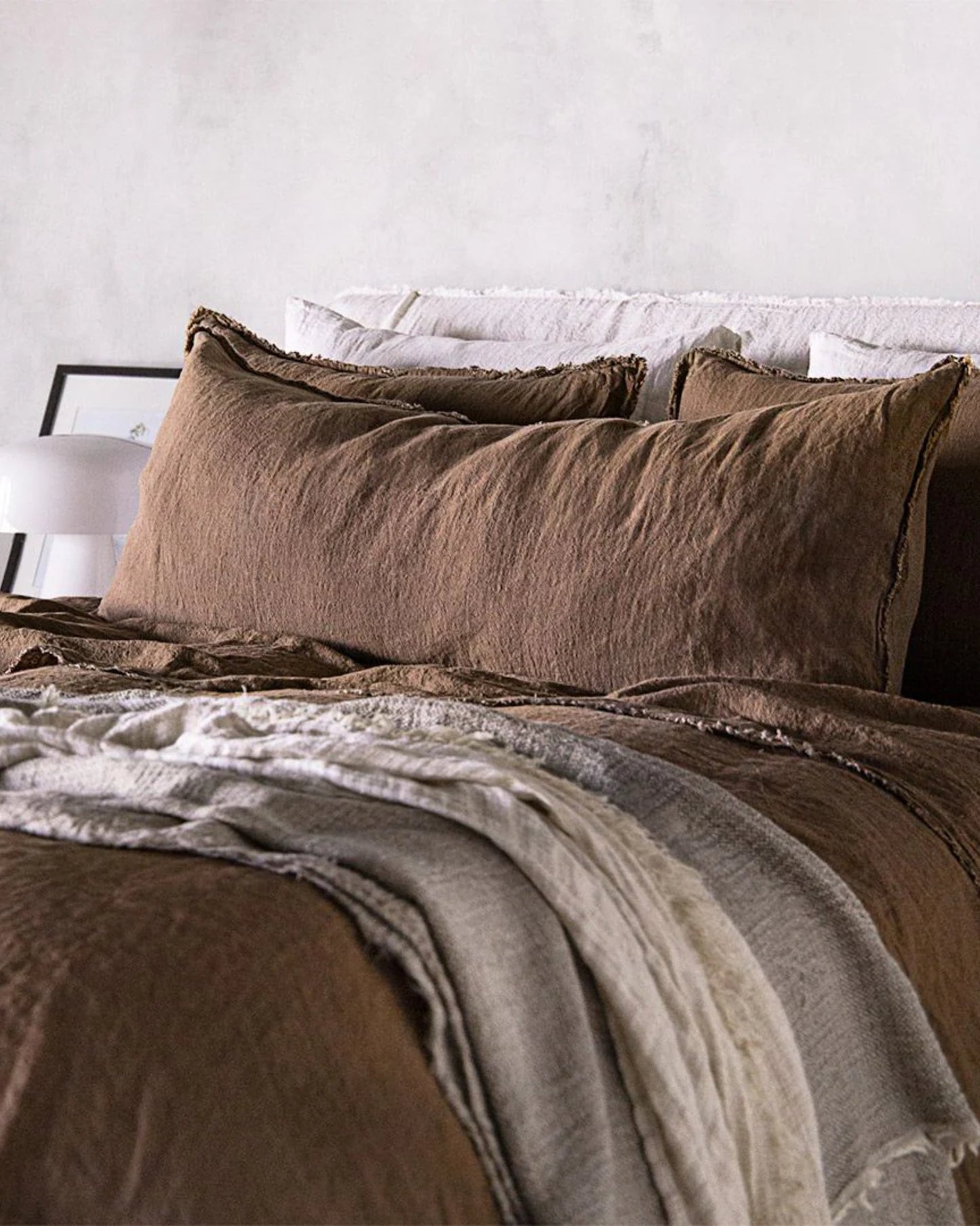 The Hale Flocca Body Pillow Case by Hale Mercantile Co adds a touch of comfort and style to a cozy bed dressed in brown and cream linen bedding. The setting includes an array of pillows, a layered blanket, and is complemented by a minimalist white lamp and framed picture against a light gray wall.