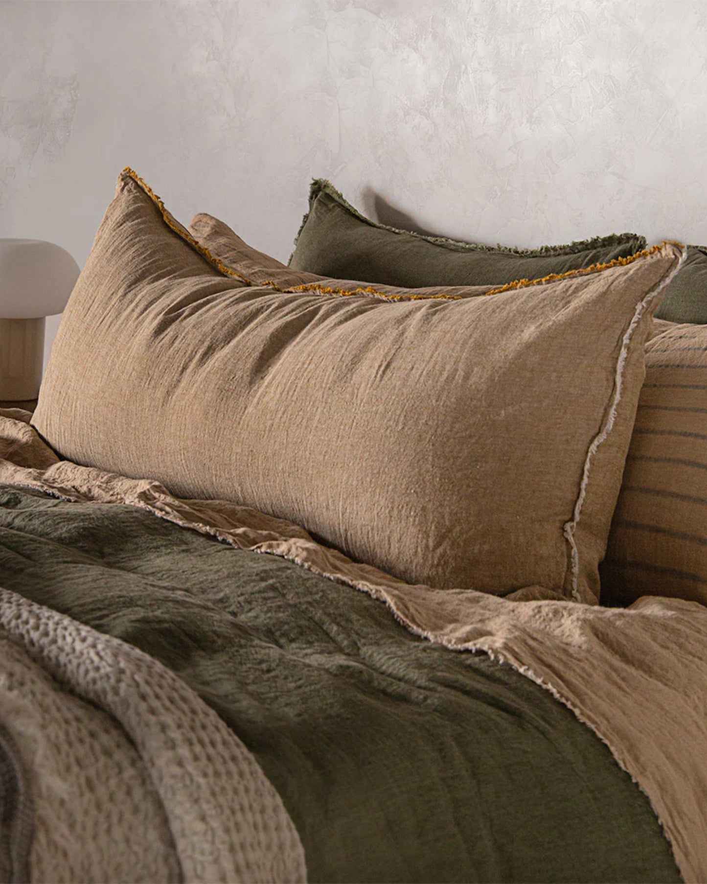 A cozy bed is adorned with layered linen bedding in earthy tones of beige and green, complemented by the Hale Mercantile Co's Flocca Body Pillow Case. This large, textured pillow case, with its charming frayed edges, rests gracefully against the headboard, creating an atmosphere that exudes warmth and comfort.