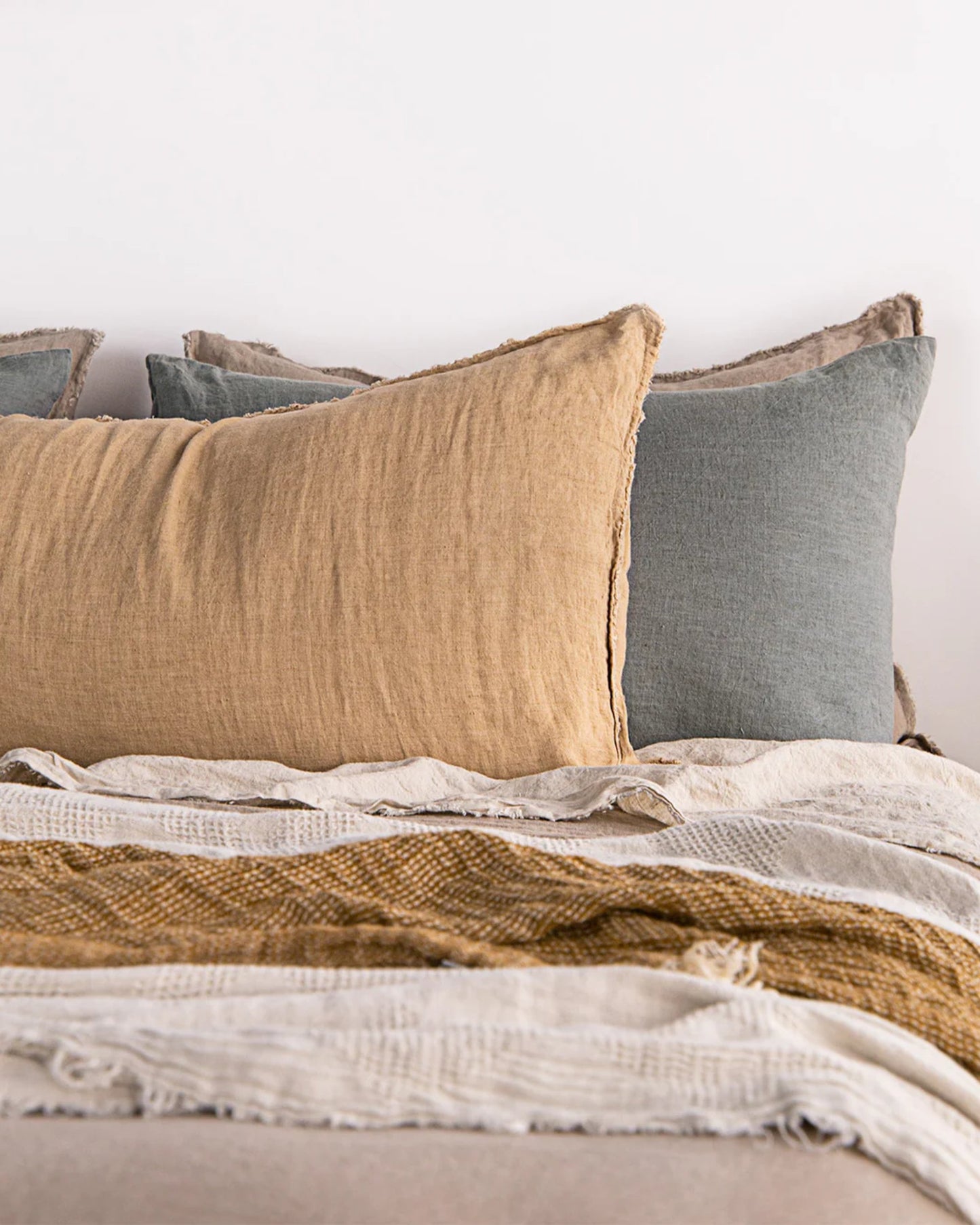 A neatly made bed featuring the Hale Flocca Body Pillow Case by Hale Mercantile Co., adorned with linen pillows in earthy tan and gray tones, complemented by layered textured blankets in warm beige and mustard hues, creates a cozy and inviting atmosphere.