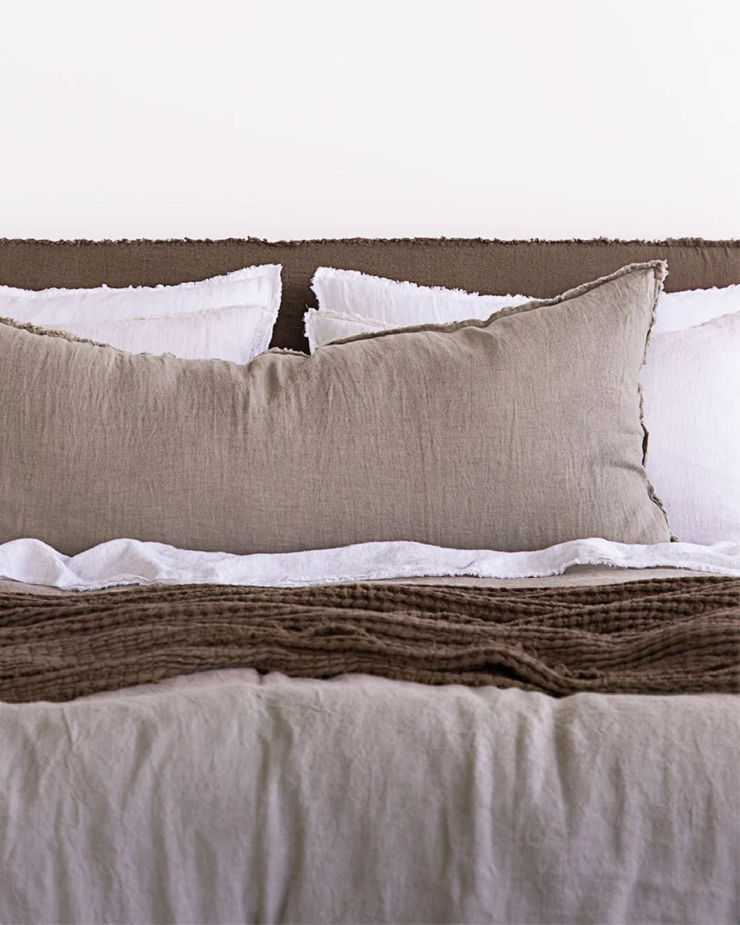 A neatly made bed with the Hale Flocca Body Pillow Case from Hale Mercantile Co adds a touch of elegance with its white and beige tones against a brown headboard. The bed is dressed in a beige duvet, complemented by a textured brown throw blanket, creating a cozy and minimalist aesthetic.