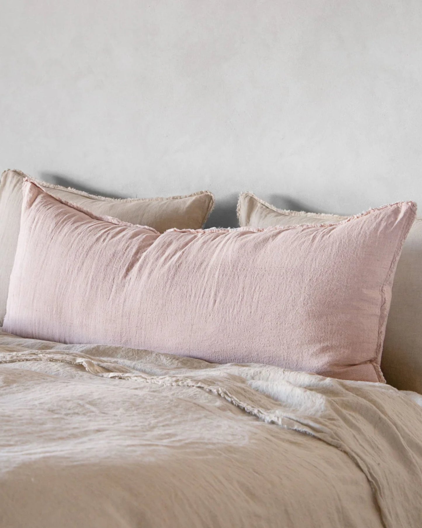 A neatly made bed showcases a large, soft pillow encased in the Hale Mercantile Co's Flocca Body Pillow Case in light pink, paired with a beige duvet. The light gray wall in the background enhances the calm and cozy atmosphere of the room.