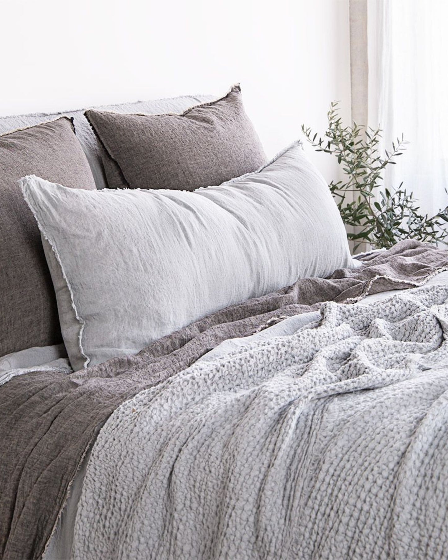 A cozy bedroom with a neatly made bed featuring textured grey and white bedding from Hale Mercantile Co. The Hale Flocca Body Pillow Case adds to the array of grey pillows and a fluffy blanket. A potted plant sits in the background by the window.